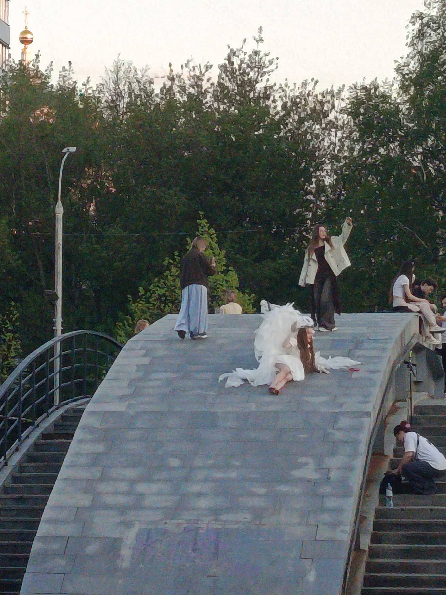 A scene shows a group of people on a stone pedestrian bridge or walkway. A woman in a white wedding dress lies prone on the walkway, while another woman in a white jacket stands with her arm raised. Other individuals sit or stand nearby, including one person squatting on the steps of the bridge. In the background, there are trees and a lamppost; in the distance, a golden dome atop a building is visible, suggesting the location is Moscow, Russia.