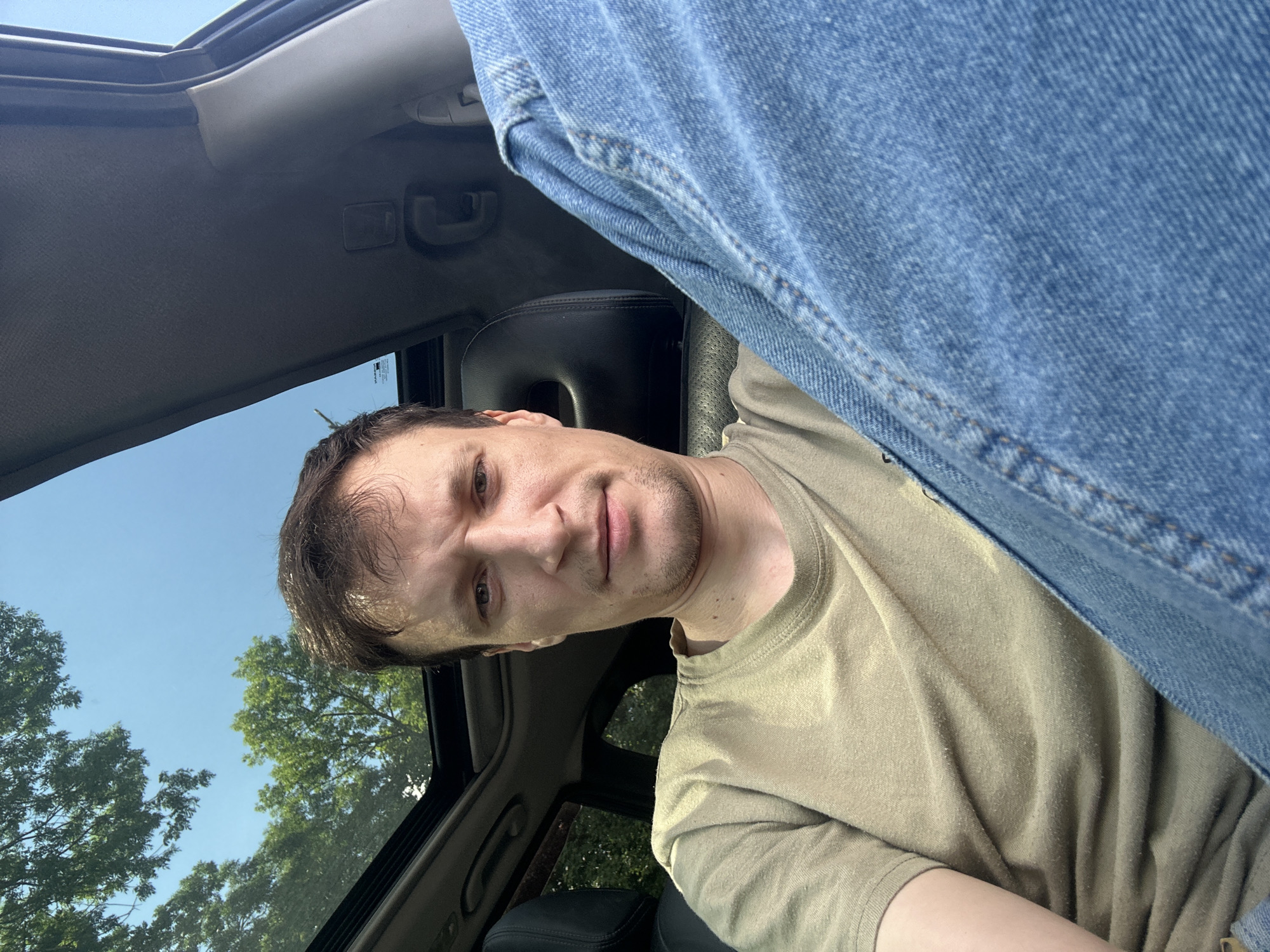A man is sitting inside a car with a sunroof open, looking directly at the camera. He is wearing a light brown t-shirt and blue jeans. The car's interior, including the headrest and part of the dashboard, is visible. Outside the car, green trees and a clear blue sky can be seen through the sunroof. The image appears to be taken from a slightly low angle, with the man's legs dominating the foreground.