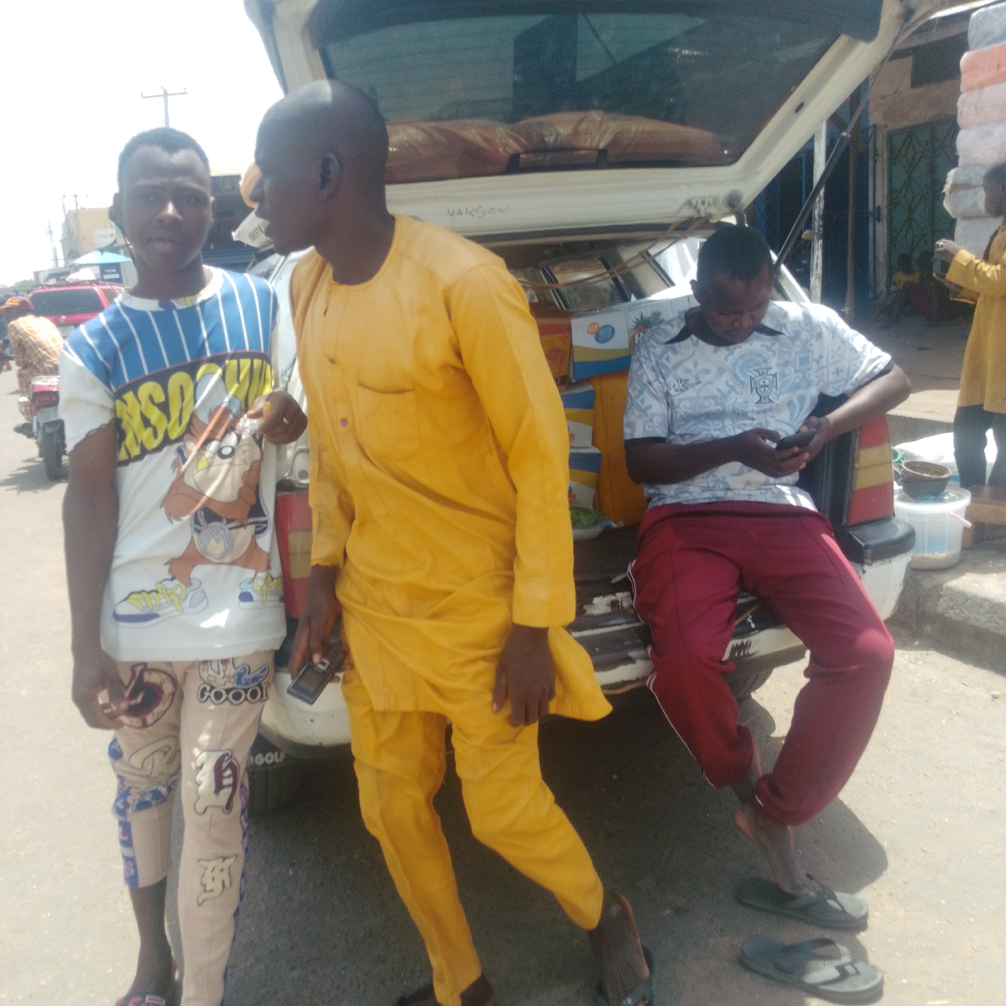 In Arawa, Nigeria, a street scene unfolds with three young men near the back of an open vehicle. The man on the left, wearing a white t-shirt with blue stripes and a graphic print of the cartoon character Taz, stands looking towards the camera. Next to him, a man in a bright yellow traditional outfit with a mandarin collar stands with his right arm bent, holding a mobile phone. To the right, another man is seated on the edge of the vehicle, wearing a light blue t-shirt with a crest and patterned design, along with maroon sweatpants. He is also looking at a mobile phone he holds in his hands. The vehicle appears to be a van or SUV, with its rear hatch open, revealing stacked items, possibly merchandise. The ground is paved with asphalt. In the background, a building with a green, patterned door is visible, and further in the distance, other individuals and structures suggest a busy marketplace or roadside commercial area. The lighting indicates a daytime scene with bright sunlight casting shadows.