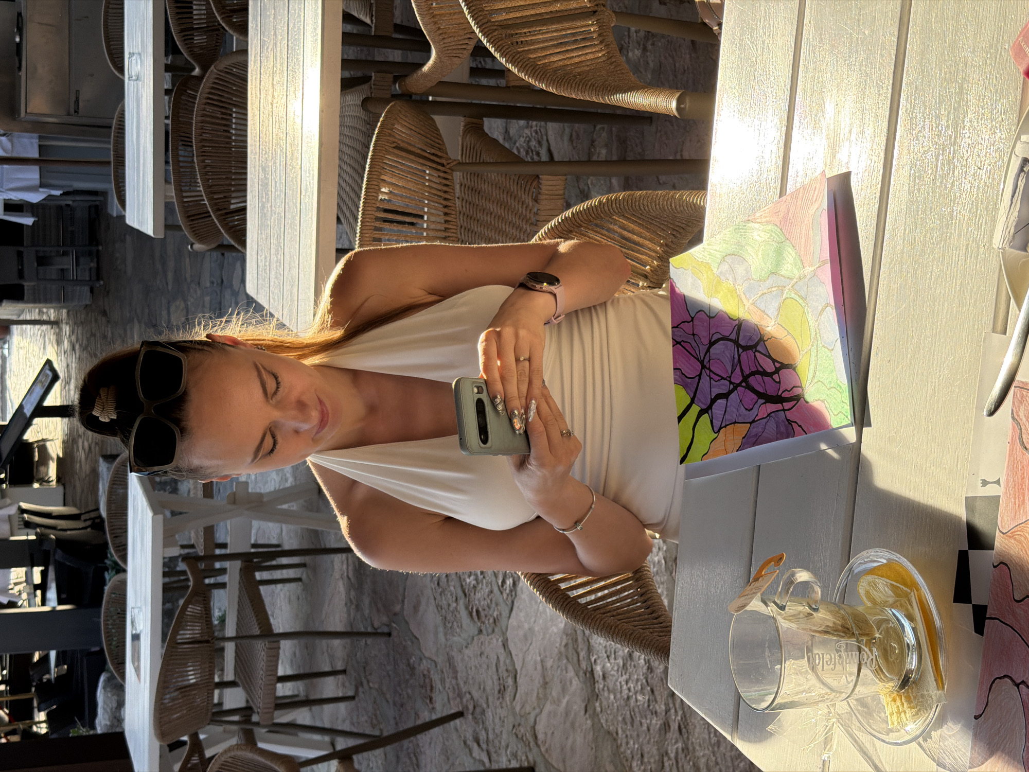 A woman is sitting at a table outside, likely in a restaurant or cafe in Budva, Montenegro. She is wearing a white halter top and sunglasses perched on her head, and she is holding a smartphone in her hands, possibly taking a photo or using it to navigate. On the table is a glass with a drink, possibly lemonade, and a colorful drawing or artwork. The surrounding area appears to be a patio or outdoor dining area with rattan chairs and stone pavement. The lighting suggests it is daytime, and there is strong sunlight present.
