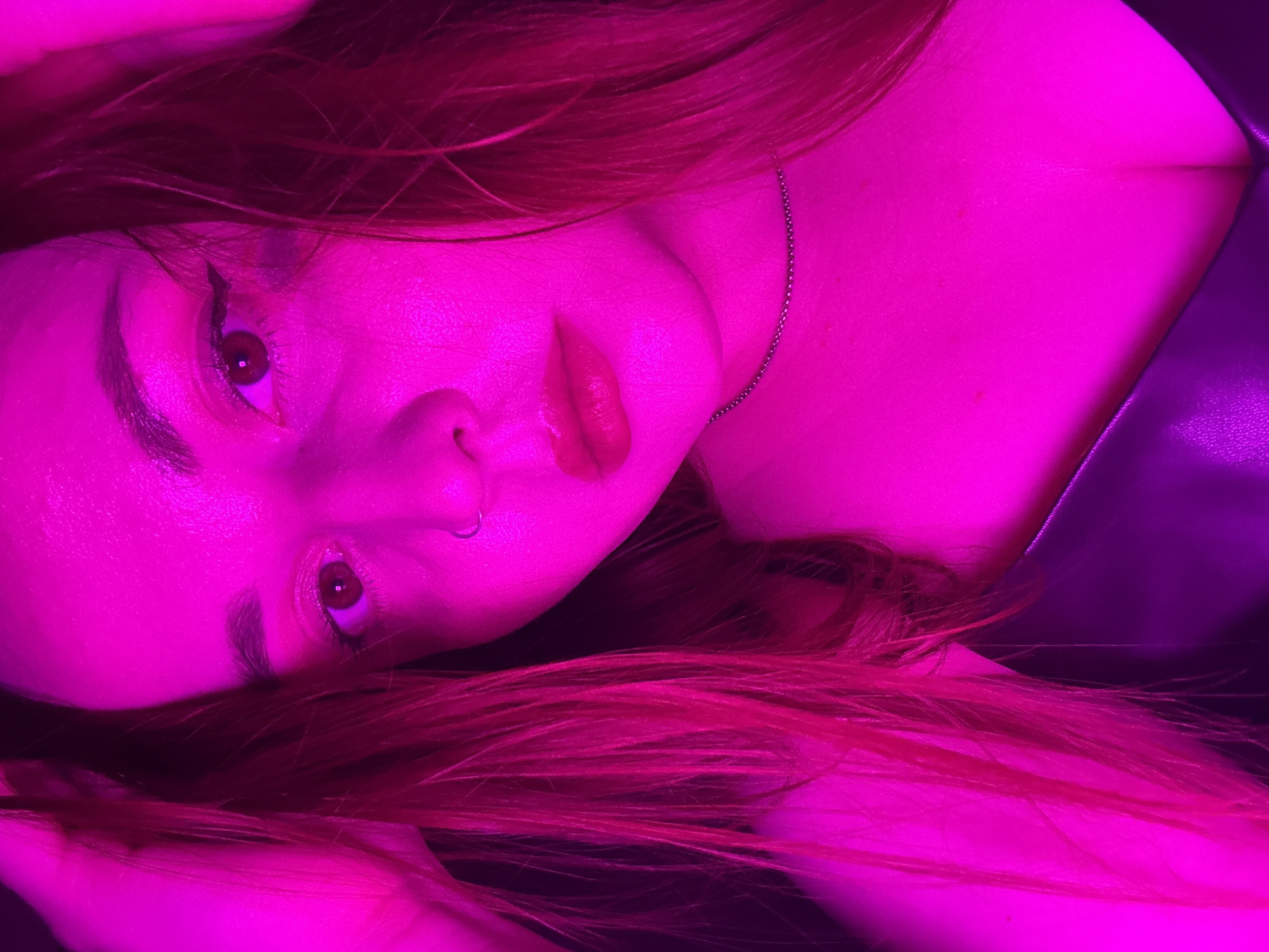 A close-up, tilted shot captures a person's face and upper chest under a pink light. The individual has long, reddish-pink hair scattered around their face and chest. Their face is turned towards the viewer. The person has dark eyeliner on and a ring in their nose. A thin chain necklace is visible on their neck. Part of a dark-colored, possibly leather, garment is seen at the top right of the frame.
I am unable to identify the location.