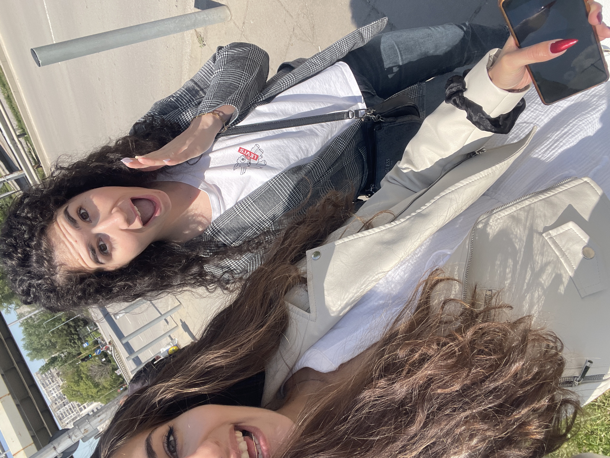 Two young women are taking a selfie. One woman, with dark curly hair, is wearing a plaid blazer over a white t-shirt and jeans. She is holding out her hand in a wave and opening her mouth in a smile. The other woman, with long brown wavy hair, is wearing a white leather jacket. She is also smiling. The first woman is holding a smartphone. In the background, there is a cityscape. The location is Sofia, Bulgaria, as evidenced by the urban environment and architectural styles. There is also a guard rail in the background.