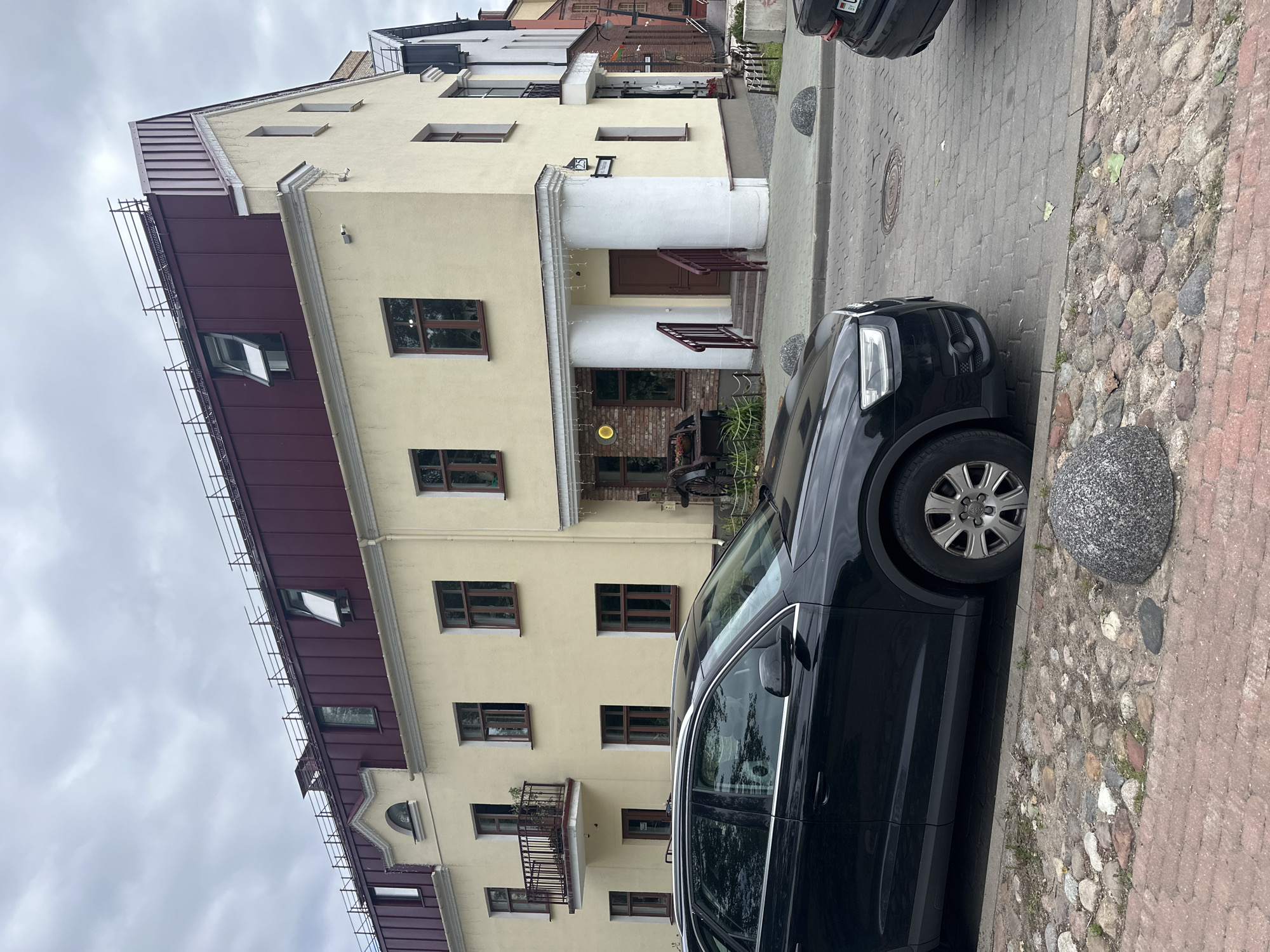 A black Audi SUV is parked on a cobblestone street in front of a light yellow building with a burgundy roof in the city of Minsk, Belarus. The building has several windows and a balcony on the second floor. There are two white columns supporting an overhang above the entrance. A black car is partially visible on the right side of the image. The sky is overcast.