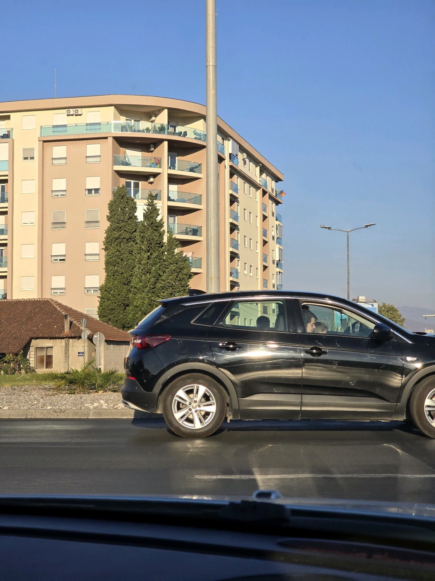 This image was taken in Podgorica, Montenegro, and captures a black SUV driving on a wet asphalt road. The car is positioned in the right foreground, with its side profile clearly visible. Inside the car, a woman and a child can be seen. The background features a tall, light-colored building with multiple balconies and windows, suggesting an urban environment. Adjacent to the building is a smaller, reddish-brown roofed structure. Tall, green trees are also present. The sky is a clear blue, indicative of a sunny day. The presence of a lamppost and what appears to be street markings further confirms an urban setting.