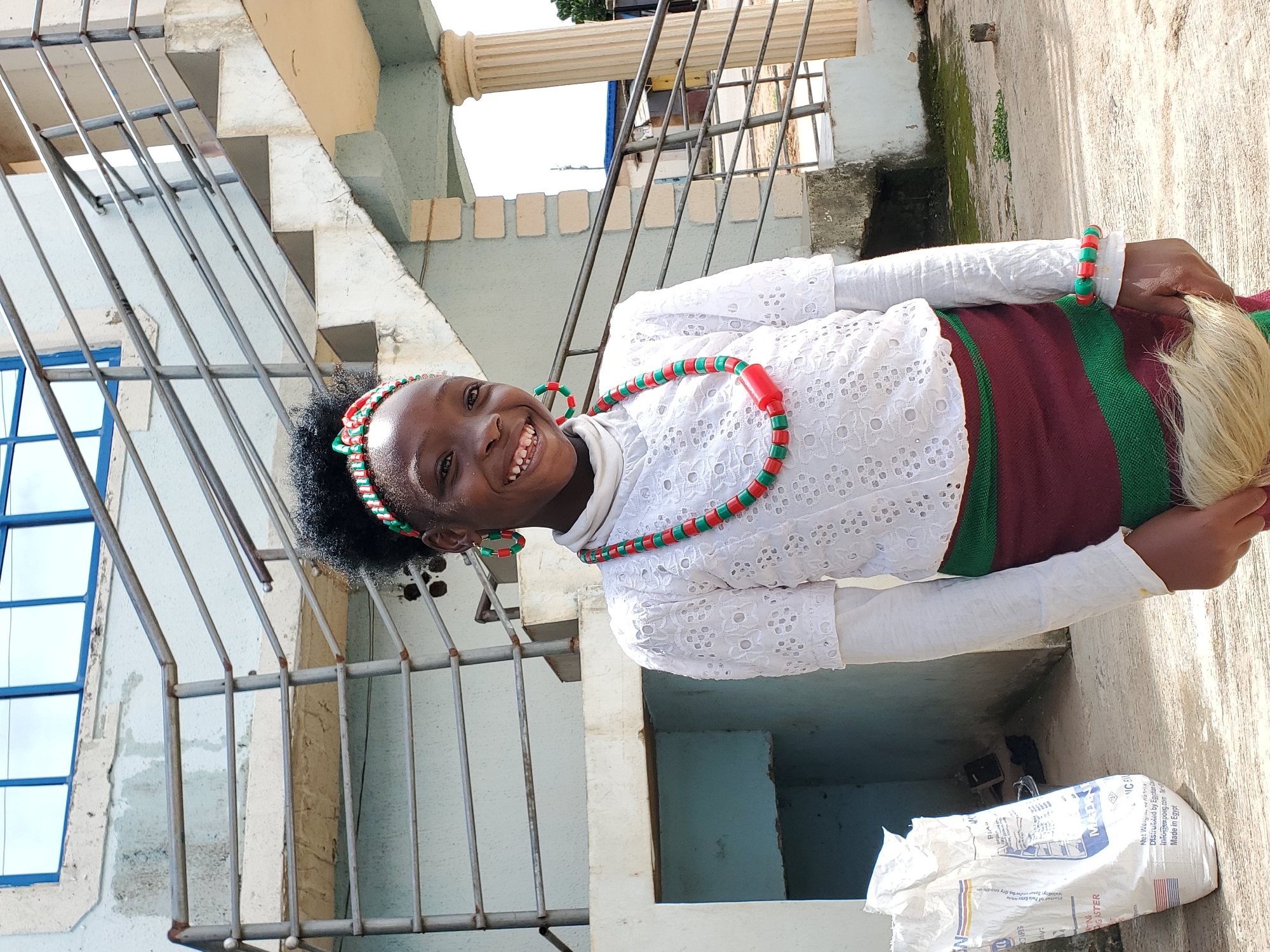 This is a vertical, medium shot of a young Black person, likely a child, posing outdoors in what appears to be a residential setting. The subject is smiling broadly and looking directly at the camera. They are wearing a white, long-sleeved top with intricate lace details on the chest and sleeves. Around their neck is a beaded necklace with alternating red and green beads, and a matching beaded bracelet can be seen on their wrist. The lower part of their attire is a skirt or fabric with horizontal stripes of maroon and green.
The background suggests an outdoor environment with architectural elements. To the left, there is a light-colored staircase with metal railings. A blue-framed window is also visible. The wall behind the subject is textured and light-colored, with some moss or green growth in places. The ground appears to be paved with concrete. A white plastic bag with some text and an American flag graphic is visible on the ground in the lower right corner. The lighting suggests it is daytime, possibly sunny, casting some shadows. The overall mood is cheerful, conveyed by the subject's wide smile.
Visible text on the bag includes "PIA," "TINE," "PIGASTER," "MADE," "Net Weight: 10 kg," "Distributed by Egyptian a," "Info@expoeg.com," and "Made in Egypt." The context provided suggests the location is Gura Top, Nigeria.
