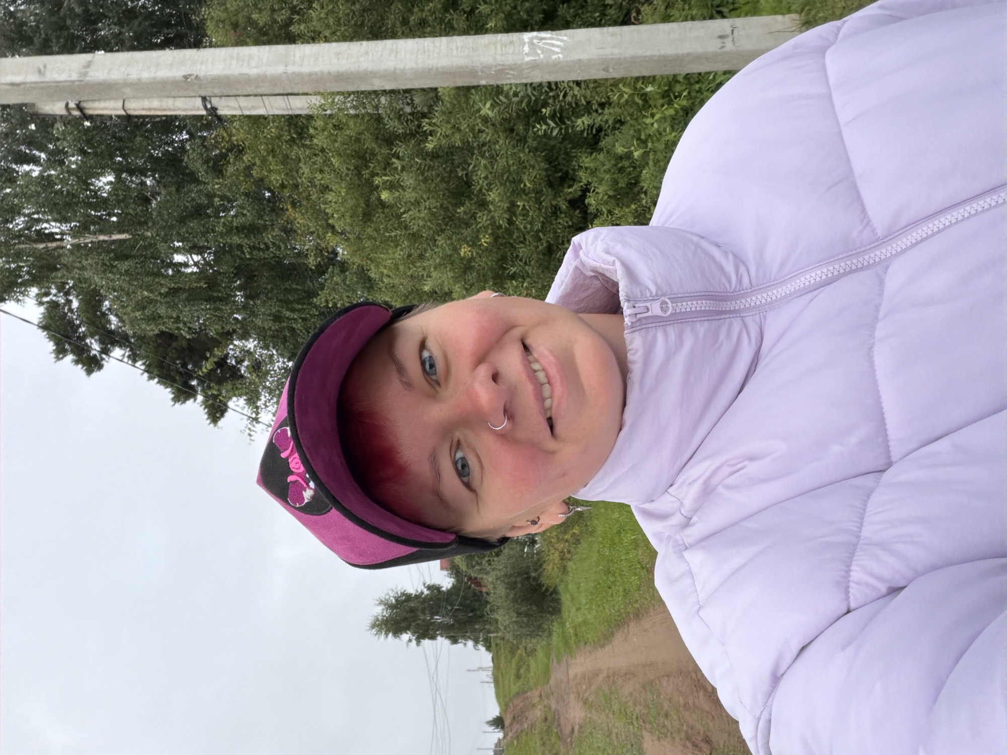 A medium shot selfie shows a fair-skinned person with short red hair, wearing a bright pink baseball cap and a light purple puffer jacket. The person has a nose ring and visible ear piercings. They are smiling at the camera. The background features a dirt road winding uphill, flanked by green grass and trees. To the right, a concrete utility pole stands beside dense green foliage and tall trees. The sky is overcast. The scene suggests an outdoor location in a rural or semi-rural area, possibly on the outskirts of Chaikovsky, Russia, given the presence of a dirt road and utility pole typical of such settings.