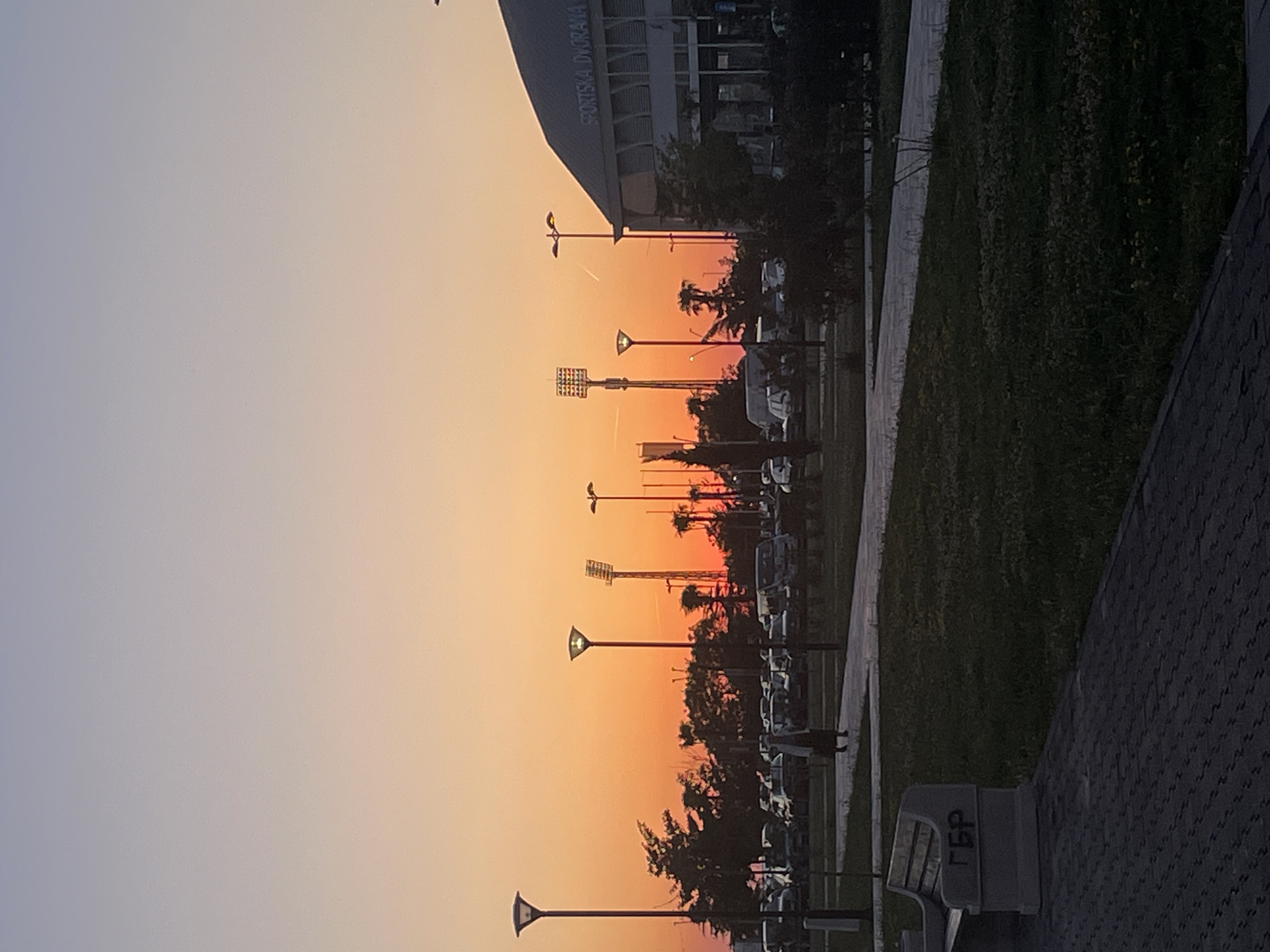 This landscape image depicts an outdoor scene at sunset in Bar, Montenegro, featuring a sports complex and adjacent public areas. The sky, covering the upper portion of the frame, transitions smoothly from a soft, hazy grey-blue at the upper left to a vivid orange and yellow glow across the horizon, indicative of late evening light. A faint airplane contrail is visible in the middle of the sky.
Silhouetted against the bright sunset are various urban elements. A large building, identified by the text "SPORTSKA DVORANA" (Sports Hall) on its facade, is prominent on the left. Numerous streetlights, some with illuminated tops, and tall stadium floodlights punctuate the mid-ground, along with dark, leafy trees and bushes that partially obscure a line of parked vehicles.
In the foreground, a paved path with a distinctive pattern runs along the right side, bordered by a strip of dark green grass. Another paved walkway is visible further to the left, with more grass beyond it. A grey park bench, featuring the Cyrillic inscription "ГБР" on its backrest, is situated in the lower right corner. The scene conveys a tranquil atmosphere as daylight fades.