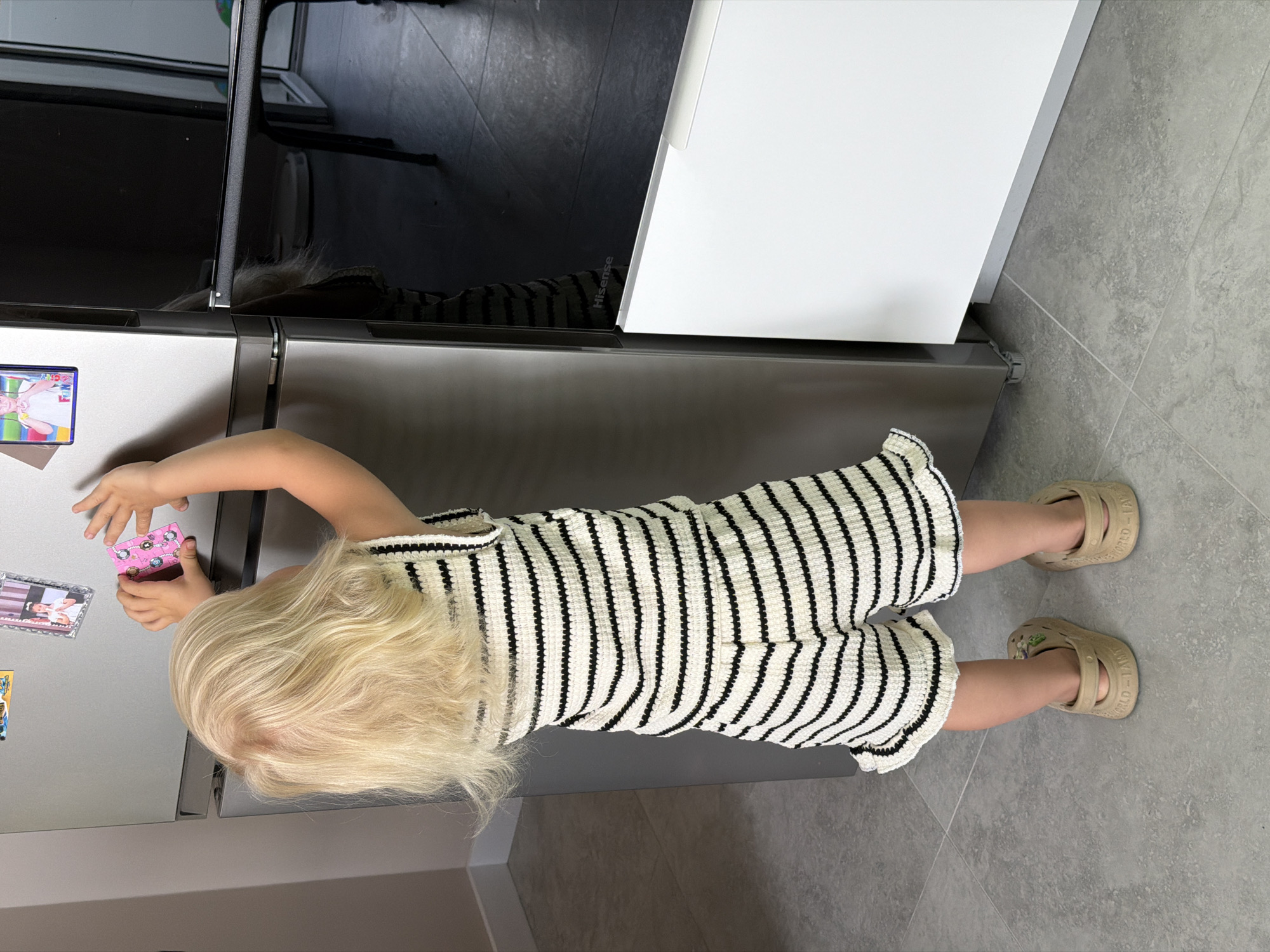 A young child with blonde hair is standing in front of a refrigerator, reaching for magnets on the door. The child is wearing a black and white striped dress and beige sandals. The refrigerator is stainless steel with a black glass door on the left side. There are several magnets on the refrigerator door, including a pink magnet with a cartoon image and a photo magnet. The floor is tiled with gray tiles. The scene appears to be indoors, possibly in a kitchen. There is no indication of the time of day or weather. The child's expression is not visible, but their posture suggests they are engaged in playing with the magnets. No visible text is discernable in the image.