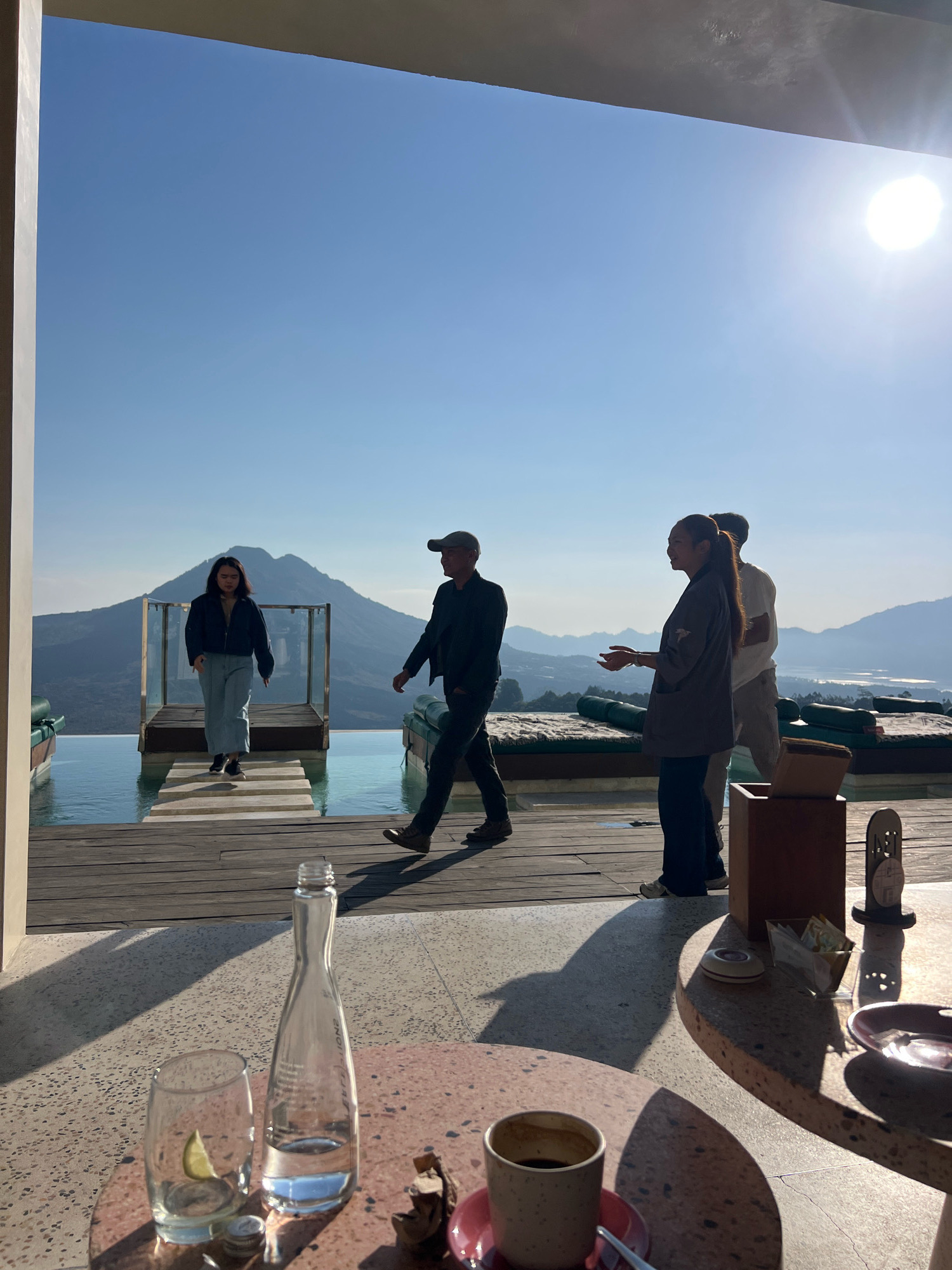 A group of four individuals are seen in the foreground and middle ground of a sunlit outdoor setting overlooking a scenic vista. In the foreground, a woman in a dark jacket and light-colored pants walks across a wooden deck towards a swimming pool. To her right, a man wearing a baseball cap and dark clothing walks away from the camera. Further back, a man and a woman stand near some lounge chairs. The woman is wearing a dark gray jacket and dark pants, and the man is wearing a white shirt and light-colored pants. They appear to be engaged in a conversation.
The background features an infinity pool with a clear blue surface, a wooden walkway extending over the pool, and lush green mountains in the distance. The sky is a clear, bright blue with the sun shining prominently, casting long shadows on the ground. The location is identified as Kintamani, Indonesia, known for its stunning views of Mount Batur and Lake Batur.
On a nearby table in the foreground, there are two glasses, one with a slice of lime, a bottle of water, and a cup of coffee on a saucer. The overall atmosphere is bright, airy, and relaxed, suggesting a vacation or leisure setting.
