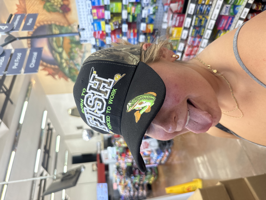 An indoor, eye-level photograph depicts a young individual, appearing female, posing within a commercial establishment in Shingle Springs, United States. The individual is positioned centrally, wearing a black and camouflage baseball cap with "BORN TO FISH" and "FORCED TO WORK" embroidered in white and green text, along with a bass fish emblem on the brim. Her face is partially obscured by the cap, and her mouth is open with her tongue fully extended. She wears a grey tank top and a gold-colored chain necklace. The background features store aisles with various products displayed on shelves. Visible overhead are fluorescent lighting fixtures, a hanging black display screen, and a department sign indicating categories such as "HABA," "Pet Food," "Charcoal," "Condiments," and "Pasta." A large wall mural depicting a natural landscape is partially visible on the far wall. A yellow wet floor sign and stacked cardboard boxes are present in the left foreground.