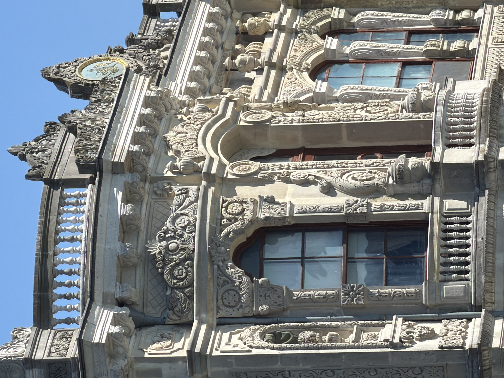 The image shows an ornate architectural detail of a building, likely a palace or a significant historical structure, in Beykoz, Turkey. The building is constructed from light-colored stone and features intricate carvings and decorative elements. Prominently visible are arched windows with dark frames, a balustrade, and elaborate friezes adorned with floral and geometric patterns. A circular medallion with Arabic calligraphy and a golden border is positioned at the top of the structure. The sky is clear and blue, suggesting fair weather. The style of the architecture appears to be a blend of Ottoman and European influences, common in Istanbul's historical buildings.