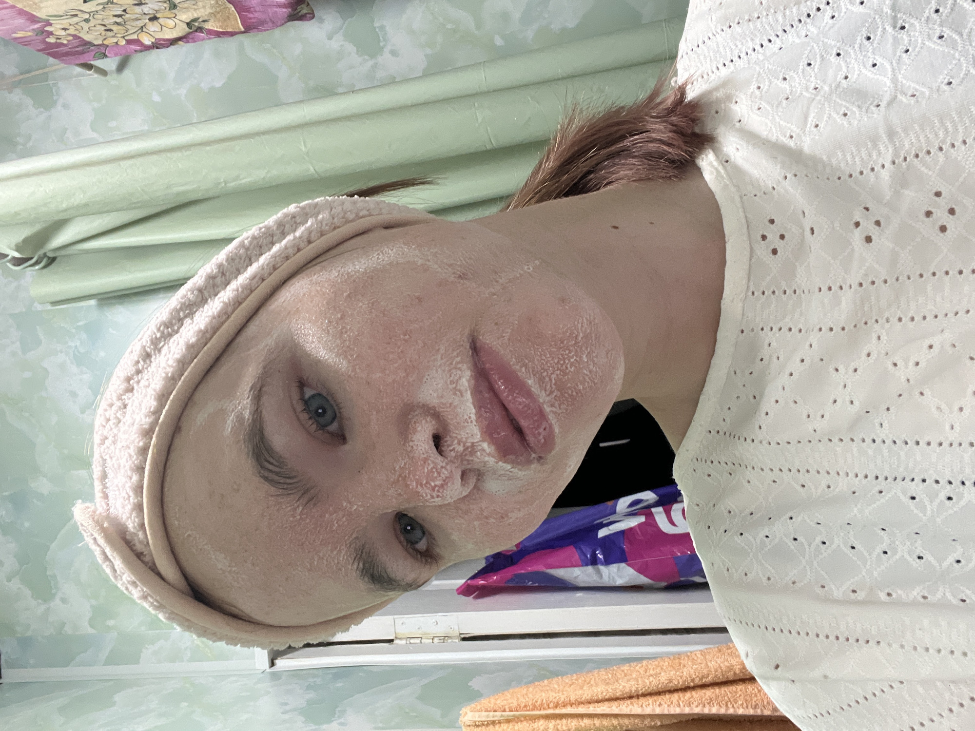 The image, rotated 90 degrees clockwise, depicts an individual engaged in a personal care routine indoors. A person with fair skin and blue eyes is shown from the chest up, their face covered in a light-colored, possibly white, facial mask. They are wearing a light-colored, possibly beige, terry cloth headband, which holds their reddish-brown hair back. A white top with an intricate eyelet or embroidered pattern is visible on their torso.
The background features a light green, possibly marbled or textured, wall or surface, along with a light-colored window blind. To the right of the person's head (when oriented upright), a purple and white plastic bag with partial, indistinct text that appears to read "SBR" is visible. Below the bag, a folded orange or peach-colored towel rests on a surface. In the top left corner of the image, a piece of floral-patterned fabric, featuring yellow flowers on a purple and pink background, is partially visible, likely a pillowcase or curtain. The overall scene suggests a domestic environment, such as a bathroom or bedroom, consistent with a self-care activity taking place in Chaikovsky, Russia. No specific time of day or outdoor weather is discernible. The person's expression is neutral as they look towards the camera.