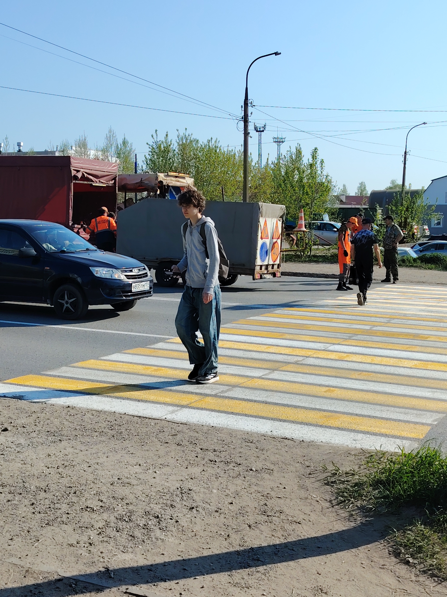 The scene is in the city of Chaikovsky, Russia. A young man with curly hair is walking across a yellow and white striped crosswalk. He is wearing a gray hooded sweatshirt, baggy blue jeans, and black sneakers, and carrying a black backpack. A black car is stopped at the crosswalk to his left. Behind the car, construction or road work is underway, with orange-vested workers visible around a maroon truck. There is a gray trailer with orange and blue warning signs attached to the back, and at least two additional workers wearing orange vests are walking across the crosswalk behind the young man. Other people and vehicles are visible in the background. The scene takes place on a bright, sunny day. Utility poles and wires are overhead. The immediate foreground shows a dirt shoulder next to the crosswalk with some grass.