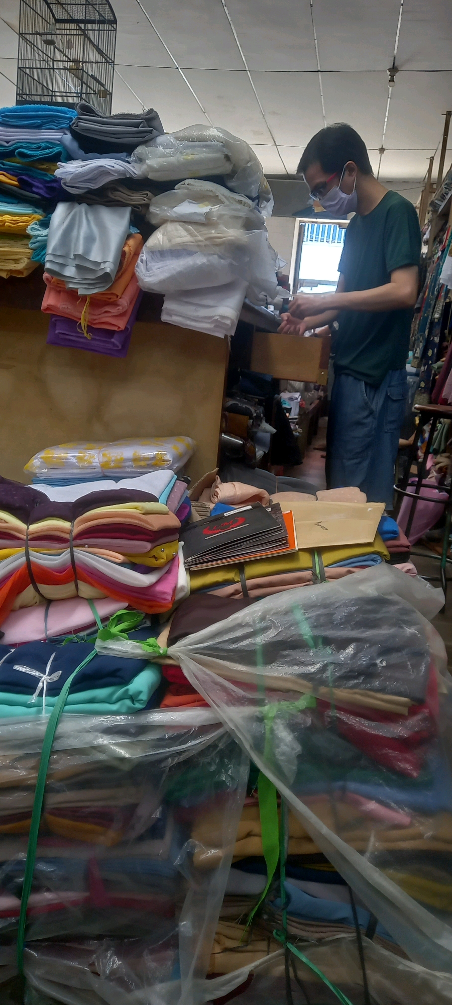 A man wearing a dark green t-shirt, blue jeans, and a face mask is standing in what appears to be a textile shop in Jember, Indonesia. He is facing towards a wooden drawer, with his hands positioned as if he is reaching into it or sorting items. The shop is filled with stacks of colorful fabrics, neatly folded and bound with strings or tape. Some fabrics are transparently wrapped. In the background, a birdcage with two birds is visible on top of a stack of fabrics. The overall impression is a well-stocked fabric store with various textiles on display and organized for sale.