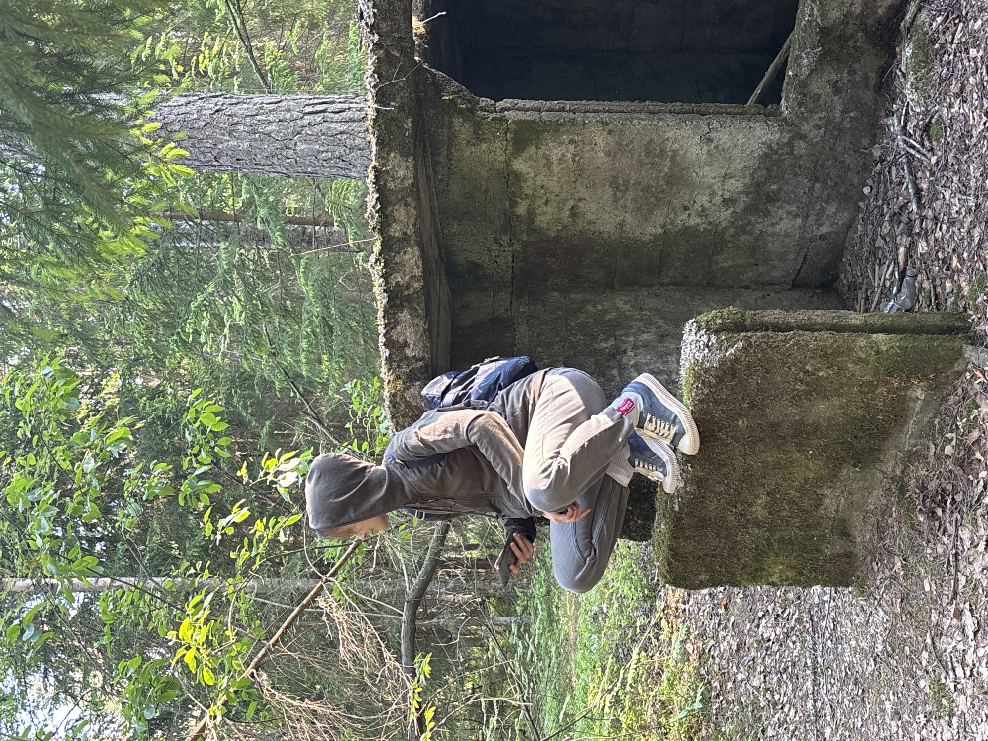 This image shows a person in a crouched position on a moss-covered concrete block within a forest. The subject, wearing a dark hooded jacket, grey pants, and blue and white sneakers, appears to be looking down at a smartphone or small object held in their hand, or into an opening of the concrete structure. A dark backpack is visible on their back.
The surrounding environment is a dense forest, with tall trees featuring both needle-like (coniferous) and broad (deciduous) leaves, indicating a mixed woodland. Bright daylight illuminates the scene, filtering through the tree canopy. The ground is covered with fallen leaves and dirt.
The concrete structure is old and weathered, significantly covered in green moss, suggesting it has been abandoned or unused for a considerable time. It appears to be part of a larger ruin or foundation, possibly a bunker, with a dark, open cavity visible above the person. The overall impression is one of exploration or discovery within a natural and possibly historical setting. No legible text is visible. This scene is set in Komarovo, Russia.