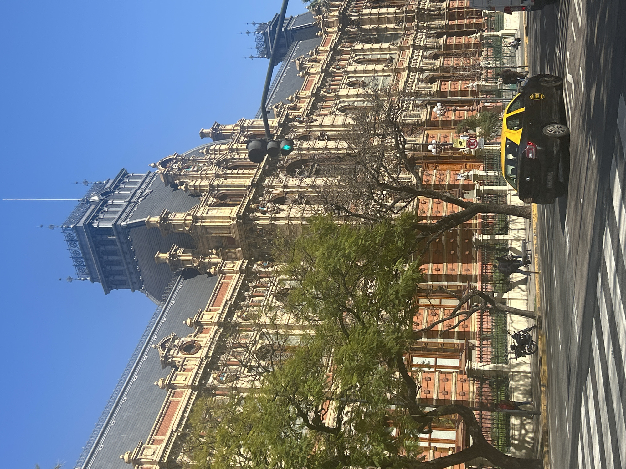 The image depicts a street scene in Buenos Aires, Argentina, dominated by a grand, ornate brick building with a dark, mansard roof and intricate stone detailing. The building exhibits multiple stories with numerous windows and decorative architectural elements, suggesting it is a significant public or institutional structure. A prominent clock tower with a spire rises from the building's roof.
In the foreground, a black taxi with a yellow roof, identifiable by the word "TAXI" on its side, is driving on the street. The taxi has license plate "XGL 159". A pedestrian crossing is visible in the lower portion of the image.
Trees line the sidewalk in front of the building, some with lush green leaves and others bare, indicating a possible seasonal change. A black motorcycle is parked on the sidewalk. Several pedestrians are present, some walking along the sidewalk, while others are near the building's entrance. A traffic light, showing a green signal, is positioned above the street. The sky is clear and blue, suggesting a sunny day. The architectural style of the building, with its rich ornamentation and mansard roof, is characteristic of late 19th or early 20th-century European-inspired architecture often found in Buenos Aires.