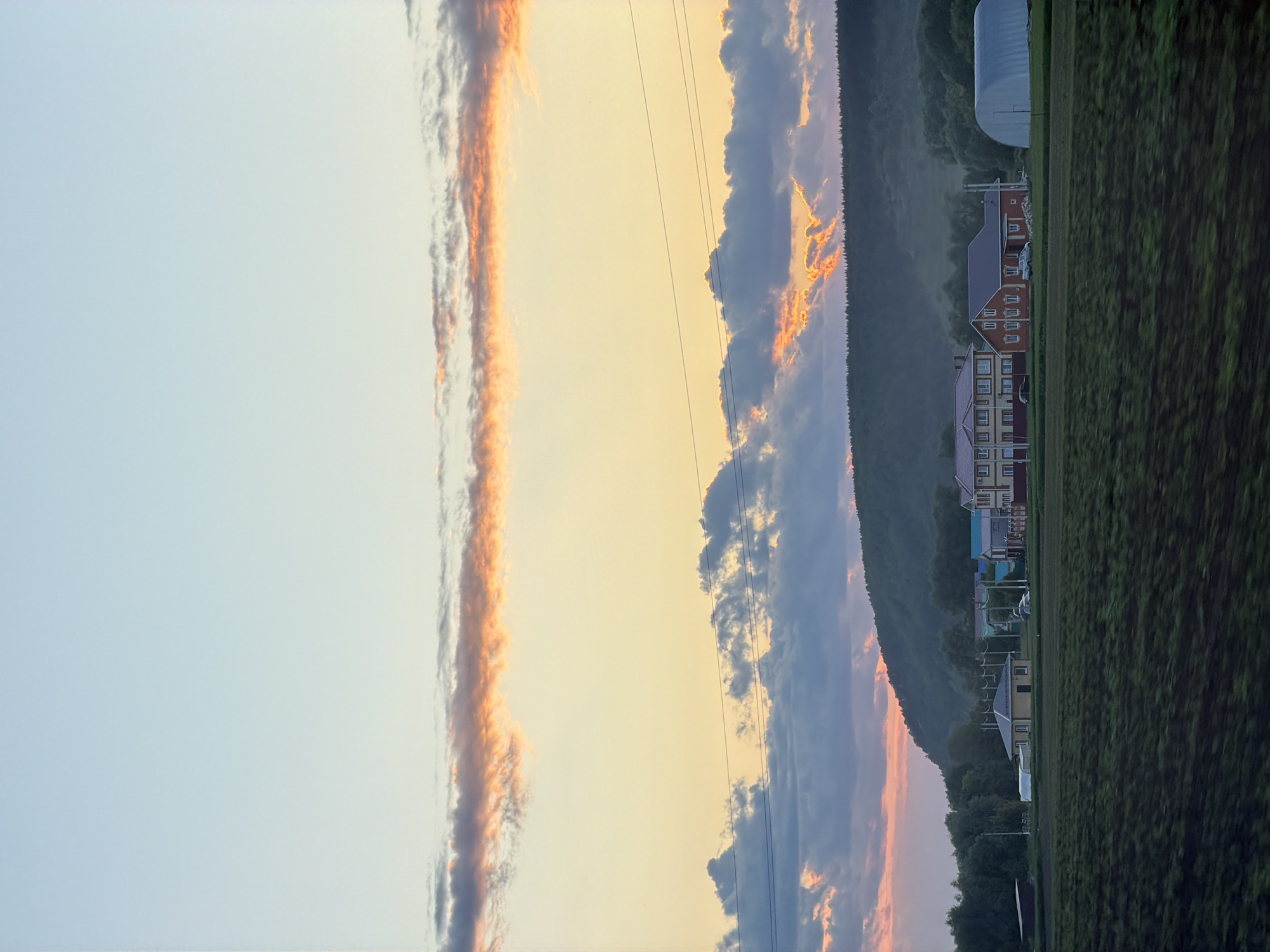This detailed description pertains to an outdoor landscape image captured during either sunrise or sunset in the city of Unknown, Unknown. The sky occupies the upper two-thirds of the frame, displaying a gradient from a pale blue at the zenith, transitioning to a warm yellow and orange above the horizon. A prominent, elongated band of stratus clouds, illuminated with orange and pink hues, extends horizontally across the upper-mid section of the sky. Below this, darker, denser cumulus clouds are present, with sections of sunlight piercing through, casting orange light on their undersides. Multiple horizontal power lines traverse the mid-sky.
The horizon is dominated by a dark, forested hill or mountain range, appearing silhouetted against the vibrant sky. Patches of mist or haze are visible at the base of the hills, particularly on the left side.
In the midground, a collection of buildings is arranged across an open field. From left to right, these include a smaller, light-colored single-story house with a dark roof, followed by a two-story house with light-colored walls and a red roof. Adjacent to this is another two-story house constructed with red brick and featuring a dark roof. A light-colored pickup truck is parked in front of the red-brick house, and a dark car is parked near the light-colored, red-roofed house. On the far right, a large, white, semi-cylindrical Quonset hut or similar storage structure is visible.
The immediate foreground consists of a dark, tilled agricultural field, showing distinct furrows indicative of recent cultivation. No individuals are depicted in the scene. The overall lighting suggests a low sun angle, characteristic of dawn or dusk, creating strong contrasts and saturated colors in the clouds.