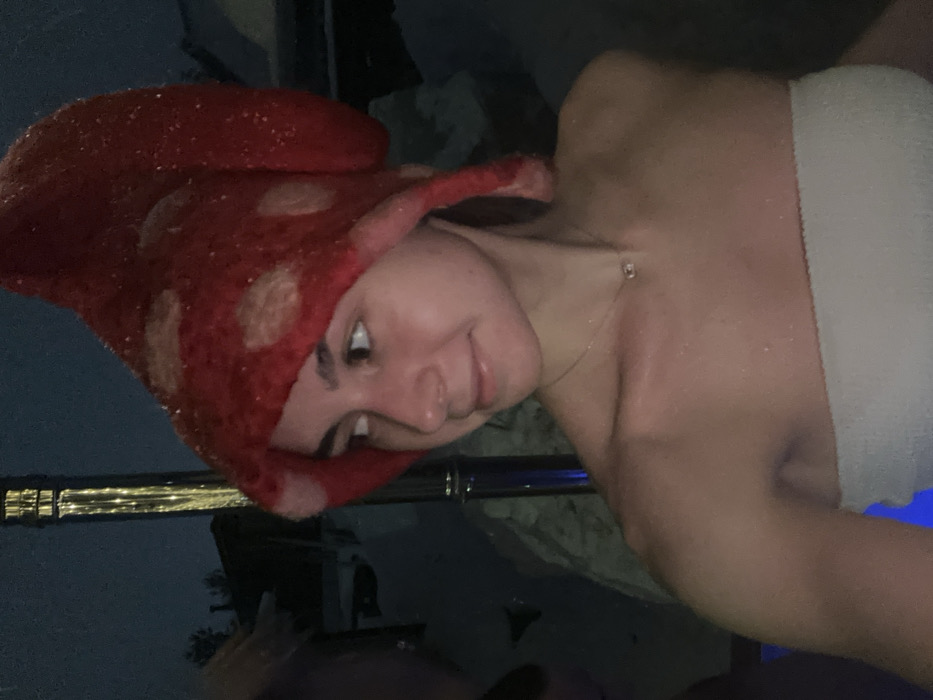 The image displays a young female outdoors at night in Базарово, Russia, likely in a hot tub or warm pool. She is captured from the chest up, facing slightly right with her head turned, looking towards the right with a subtle smile. She wears a red, felted, mushroom-shaped hat with lighter, circular spots and a white bandeau-style top. Her skin appears wet and glistens with water droplets. A delicate silver-colored necklace with a small rectangular pendant is visible around her neck. The background is dark, revealing snow-covered ground and faint outlines of structures. A tall, reflective pole, possibly a heater or light fixture, is visible behind her, emitting small points of yellow light. A blurred, dark mass, potentially another individual or object, is partially visible on the far left. The scene suggests a leisure activity in a cold, outdoor, winter environment.
