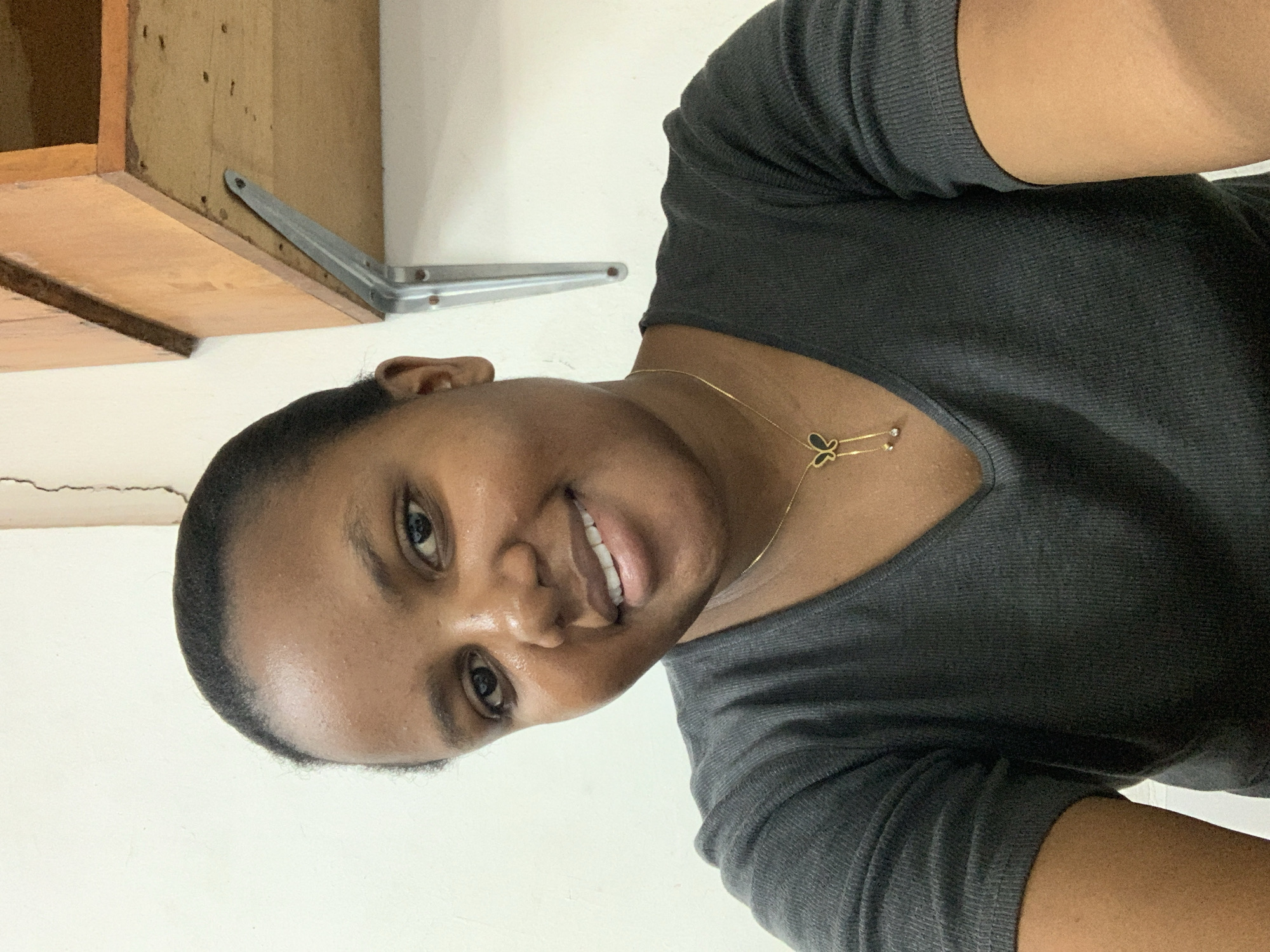 The image displays a head-and-shoulders portrait of an adult female with dark skin and dark hair, pulled back from her face. She is looking directly at the camera with a frontal gaze and a visible smile, revealing white teeth. She wears a dark grey, ribbed, short-sleeved V-neck top. Around her neck is a thin gold-colored chain with a butterfly-shaped pendant, also appearing gold-colored with dark inserts, and two small, dangling, circular elements.
The background consists of a plain, light-colored wall, possibly white or off-white. A vertical, dark crack is visible in the upper left portion of the wall. In the upper right background, a light brown wooden shelf or cabinet is visible, supported by a metallic, L-shaped bracket affixed to the wall. The underside of the wooden object shows multiple small dark marks, consistent with fastener impressions.
Based on the visual information alone, the specific location within the city of Dar es-Salaam, Tanzania, cannot be determined. The scene appears to be an indoor setting.