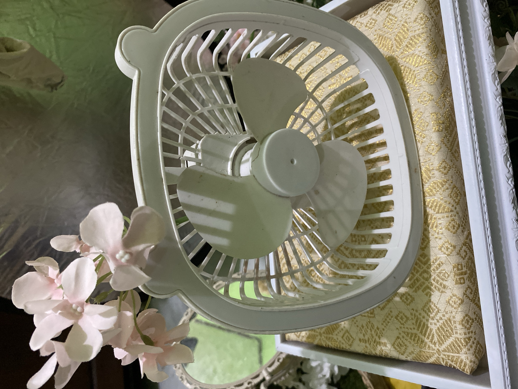 The image shows a close-up of a white, small electric fan placed on a decorative white frame. The fan is positioned in the foreground, partially obscuring a piece of fabric with an intricate gold pattern. In the lower left corner of the image, a cluster of delicate pink artificial flowers is visible, adding a soft touch to the composition. The background is blurred and suggests a domestic setting with reflective surfaces and hints of green. The lighting appears to be artificial, possibly from an indoor light source, creating subtle shadows and highlights on the fan and the surrounding objects. There is no visible text in the image. The overall mood is still and decorative, possibly part of a display or arrangement. Given the location context of Kuala Pembuang, Indonesia, this might be an item used for personal comfort in a warm climate or part of a celebratory decoration.