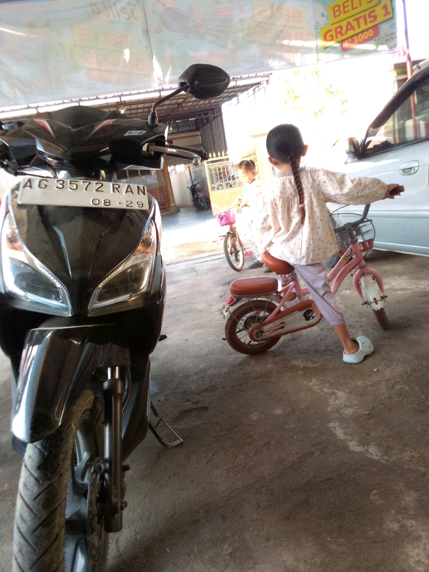 In Simo, Indonesia, a young girl with braided hair, wearing a patterned long-sleeved shirt and light purple pants, is seated on a small pink bicycle. She is looking towards the right of the frame, with her right hand on the handlebar. Beside her, to the left, is a black motorcycle with a white license plate reading "AG 3572 RAN 08-29". The motorcycle is parked. In the background, another young girl is visible near a pink bicycle. A silver car is parked on the right. The ground is a concrete surface. A banner with text is visible hanging in the upper portion of the image, and part of a building structure is present.