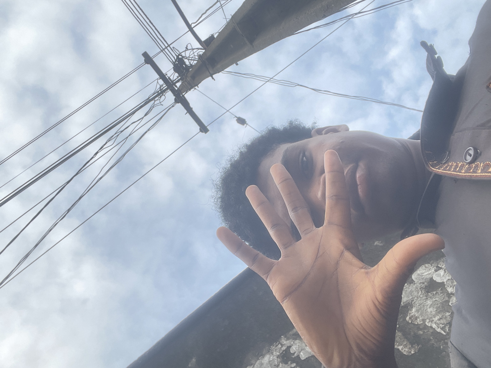 A low-angle, outdoor, eye-level medium shot captures a person holding their left hand up to cover the right side of their face. The individual has dark, curly hair and is wearing a dark jacket with ornate gold trim and a button. The background features a cloudy sky with numerous electrical wires and a utility pole on the right. A dark, textured wall runs along the left side of the frame. The scene appears to be in a city setting, possibly in Ado, Nigeria, given the presence of overhead electrical infrastructure commonly found in urban environments. The lighting suggests it is daytime.