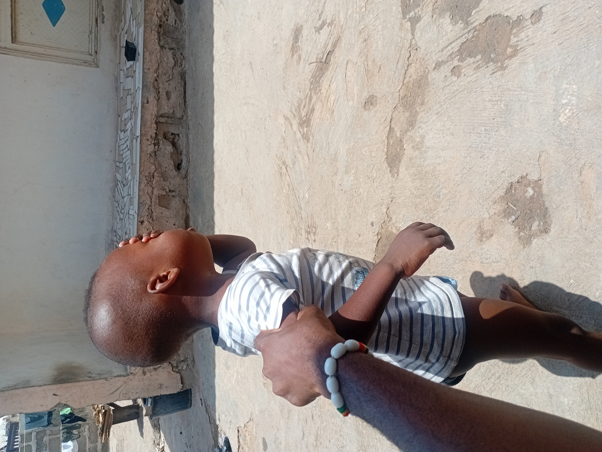 An outdoor, eye-level shot captures a young child being gently held by an adult's arm, bathed in bright sunlight. The child, with dark skin and a close-cropped haircut, wears a white t-shirt with grey stripes and blue shorts. They are looking upwards and slightly to their right, away from the camera.
The adult's dark-skinned arm is visible in the lower right, holding the child's upper left arm with their right hand. The adult wears a distinct beaded bracelet on their wrist, featuring alternating white, red, yellow, and green beads.
The setting appears to be a paved or concrete outdoor area, with irregular textures and some light-colored debris on the ground. Strong shadows cast by the subjects and objects indicate clear, sunny conditions, suggesting it is daytime. In the background, to the left, is a light-colored wall or building exterior, with some architectural details including a blue diamond shape on a panel and textured or mosaic-like elements along a vertical edge. Further back, objects resembling laundry or household items are visible on a lower ledge. The general environment is consistent with a warm climate, specifically in Lokoja, Nigeria. There is no visible text in the image.