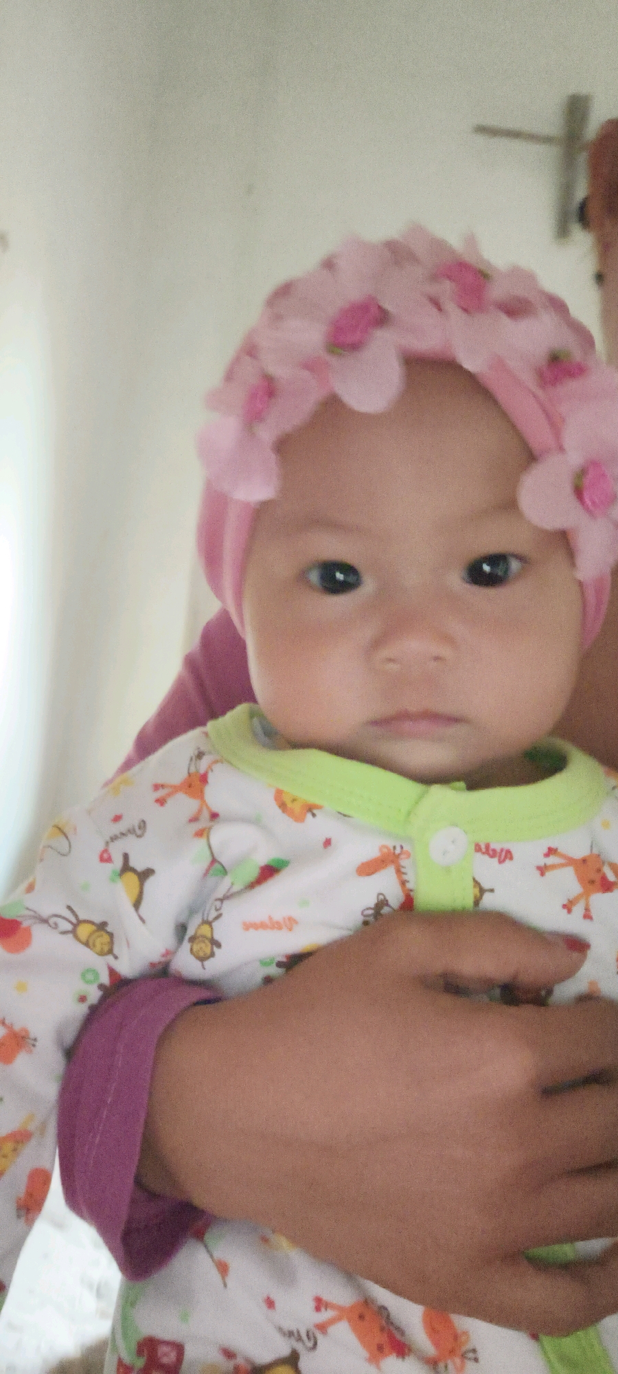The image shows a close-up of a baby wearing a pink headband with fabric flowers. The baby is being held by an adult whose arms and hands are visible. The baby is wearing a white onesie with a cartoon animal print. The background is out of focus and appears to be an indoor setting with a light-colored wall. The location of this scene in Waisai, Indonesia is not discernable from the provided image.