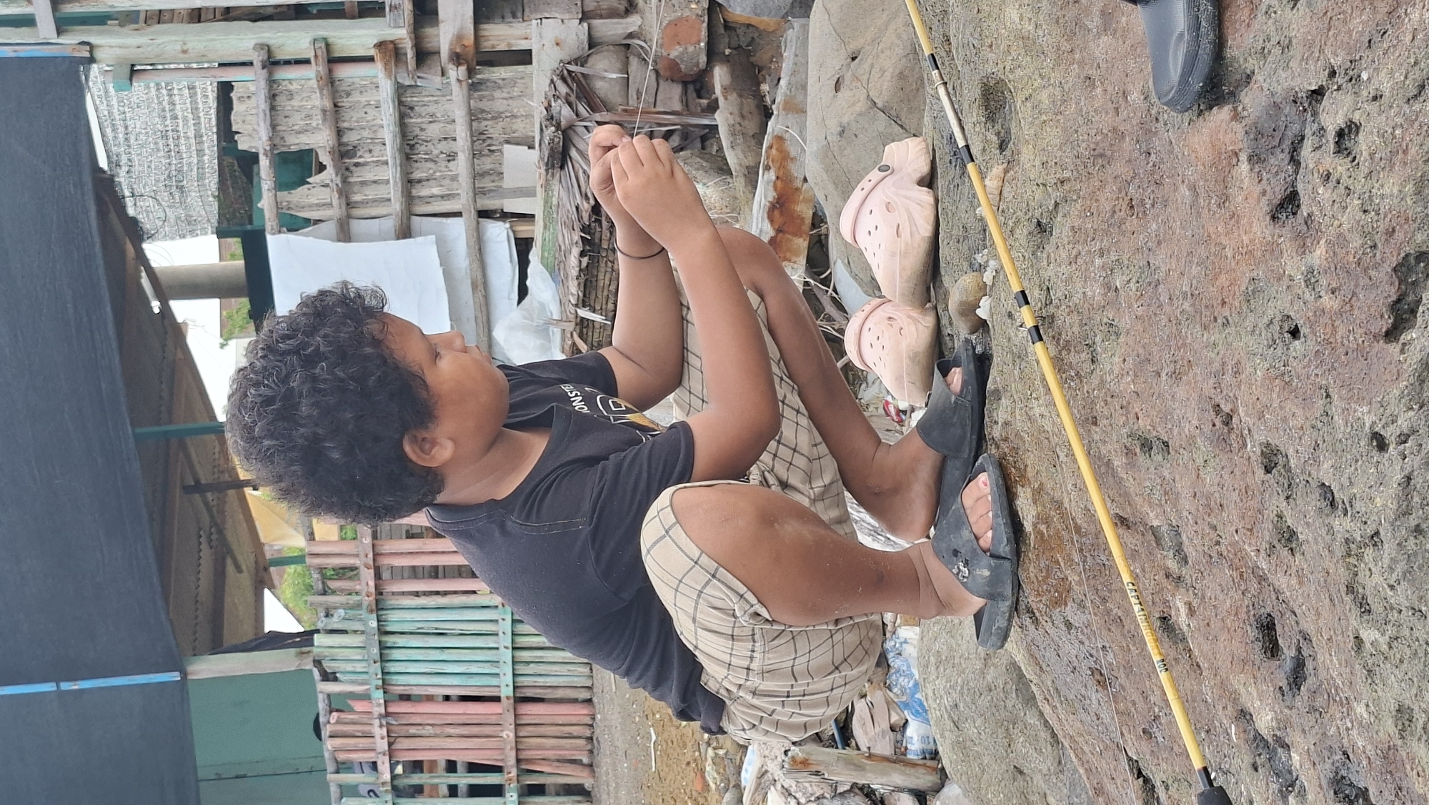 A young boy with curly dark hair is squatting on rocks near the water, holding a fishing rod. He is wearing a black t-shirt and plaid shorts, with black flip-flops on his feet. Two light pink clogs are next to him. In the background, there is a weathered wooden fence and a makeshift shelter. The location appears to be a coastal area in Lhokseumawe, Indonesia, likely a fishing spot or informal settlement.