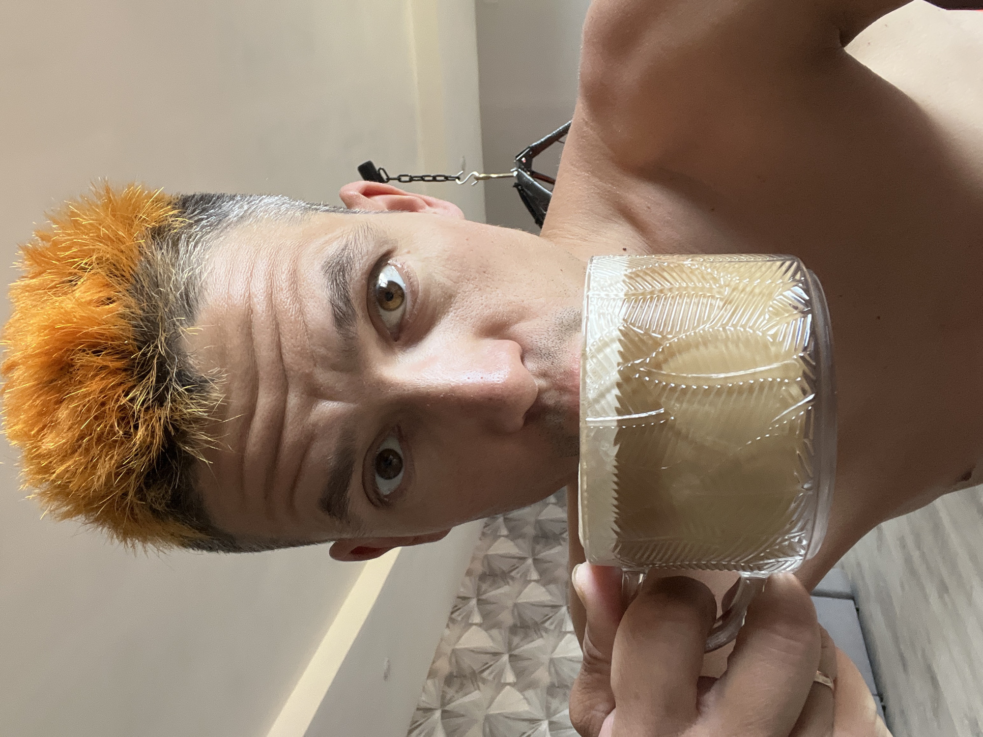 A shirtless individual with dyed orange hair and a surprised expression is holding a container of what appears to be eggs in front of their face. The person has visible lines on their forehead and light brown eyes. In the background, there is a wall with a triangular pattern and a chain visible above the left ear of the individual. The location is not identifiable as Chaikovsky, Russia from the visual information provided.