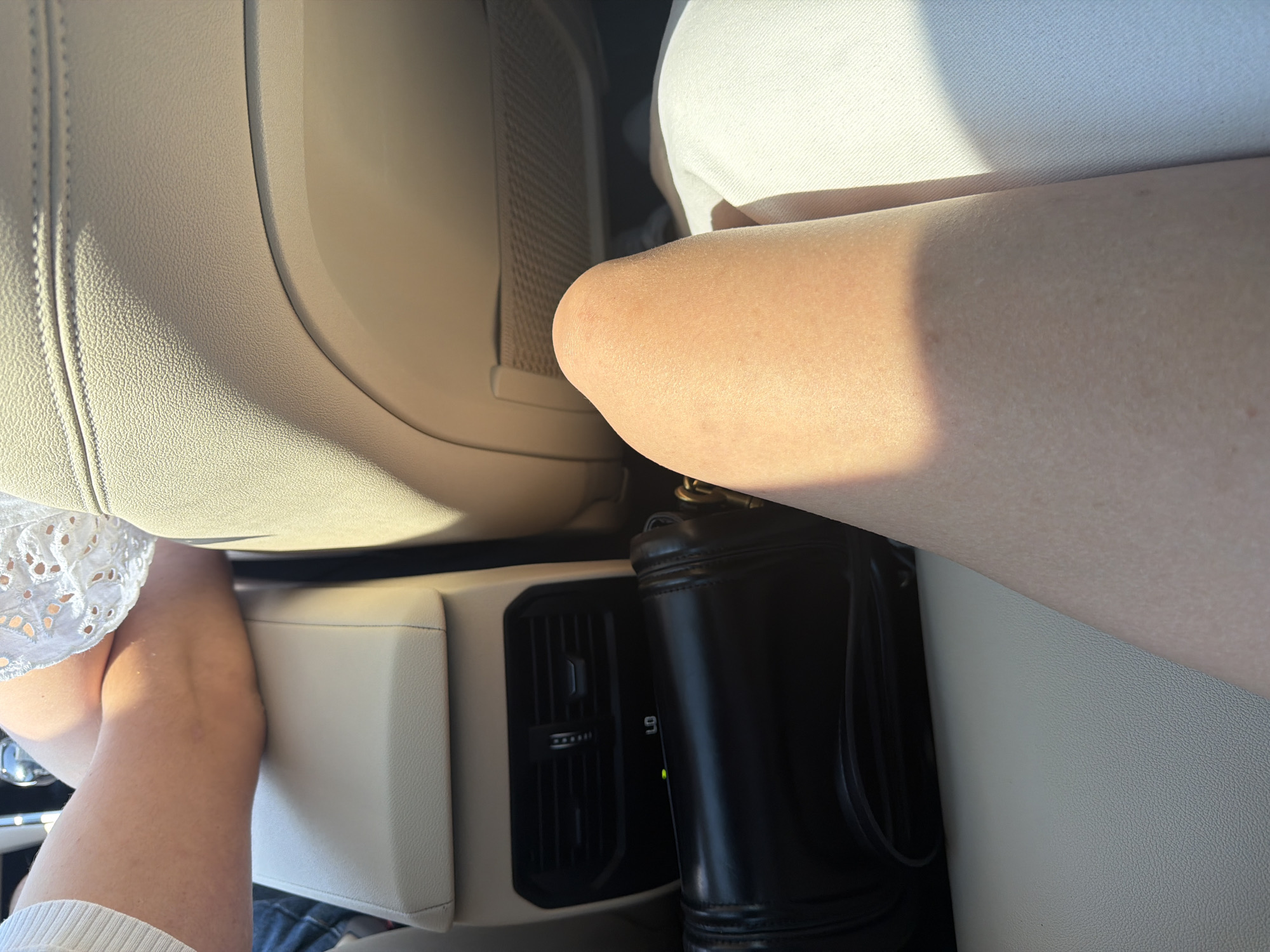 The image depicts the interior of a vehicle, likely a car, seen from the perspective of a rear passenger. The focus is on the lower body of a person, specifically their leg and knee, which occupies the foreground and right side of the frame. The leg is bare and shows a light skin tone with some minor blemishes. The person is wearing white pants, visible on the right.
In the mid-ground, a black handbag is placed on the central console or armrest of the car. Behind the handbag, a rear air vent with adjustable louvers is visible. The surrounding upholstery of the car's interior is a light, neutral color, appearing to be leather or a similar material, with visible stitching on the seatback. Sunlight streams into the car, casting shadows and highlighting the textures of the surfaces.
The image does not provide any visual cues that would allow for identification of the specific location within Chișinău, Moldova, or any other city. The scene is confined to the interior of a vehicle and offers no external environmental context.
