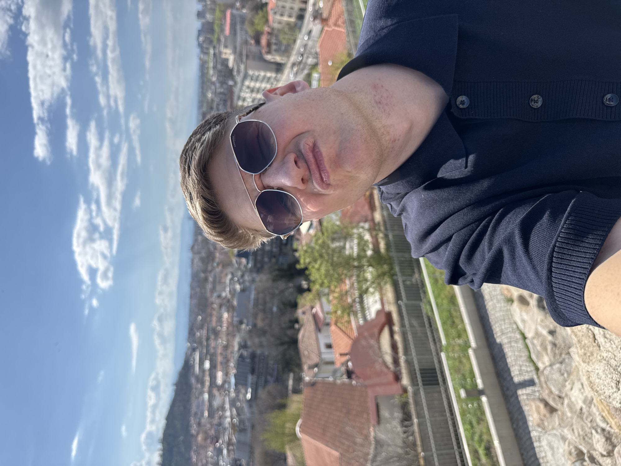 A man is the central focus, positioned in the foreground of a cityscape. He is wearing sunglasses and a dark blue button-down sweater. The background features a sprawling cityscape with a view of Plovdiv, Bulgaria. The foreground shows a stone and metal railing suggesting an elevated viewpoint, possibly a balcony or observation area. The sky is visible with some cloud cover. Buildings with red-tiled roofs and greenery are visible below.