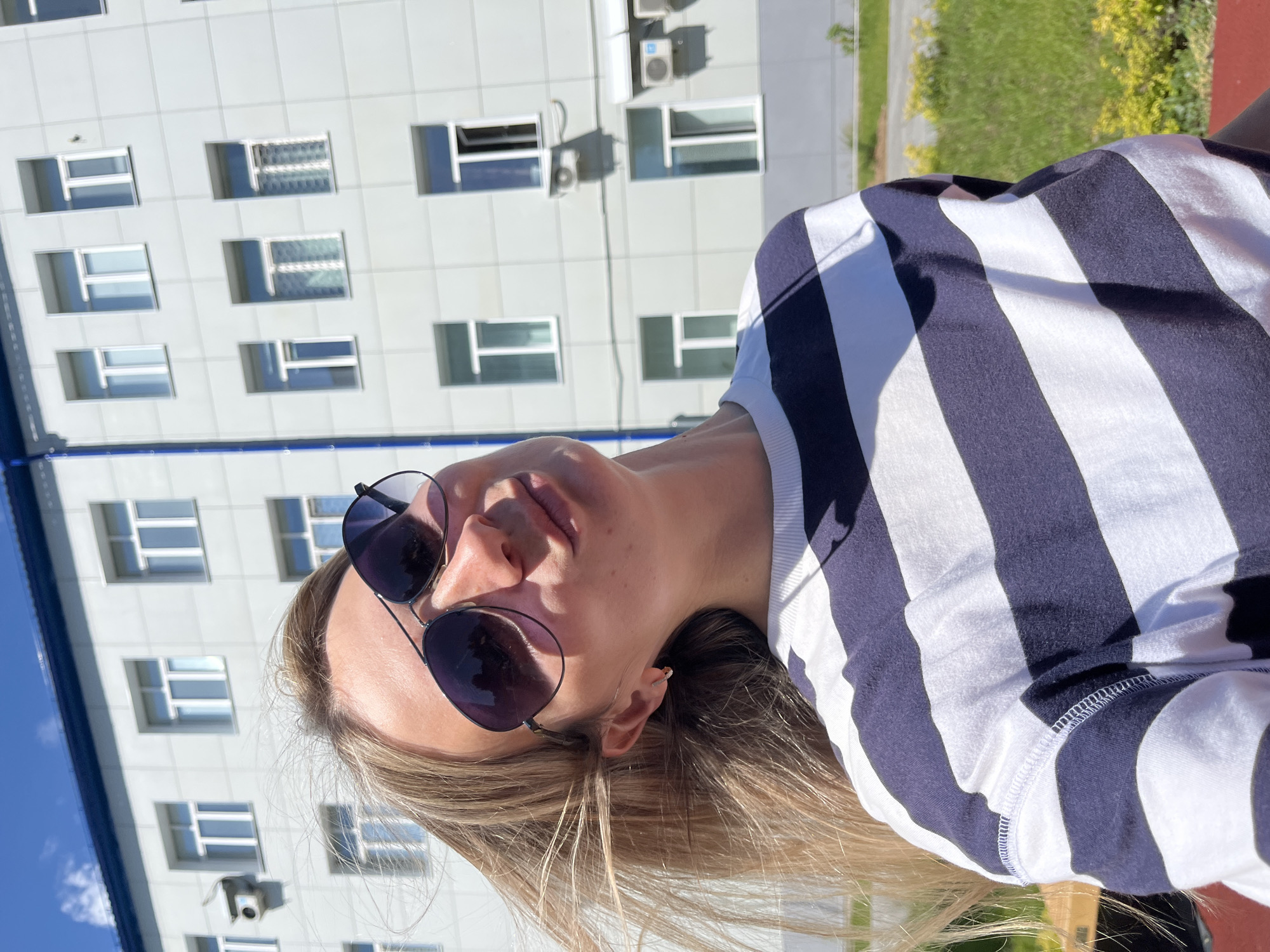 A woman is taking a selfie outdoors, with a modern building in the background. She is wearing sunglasses and a blue and white striped long-sleeved shirt. Her blonde hair is visible, falling around her face and shoulders. The building behind her has a grid of identical rectangular windows. The sky is a clear blue, and the sun is shining brightly, casting shadows on the building and illuminating the woman. The overall impression is that of a sunny day. The location is specified as Chaikovsky, Russia.