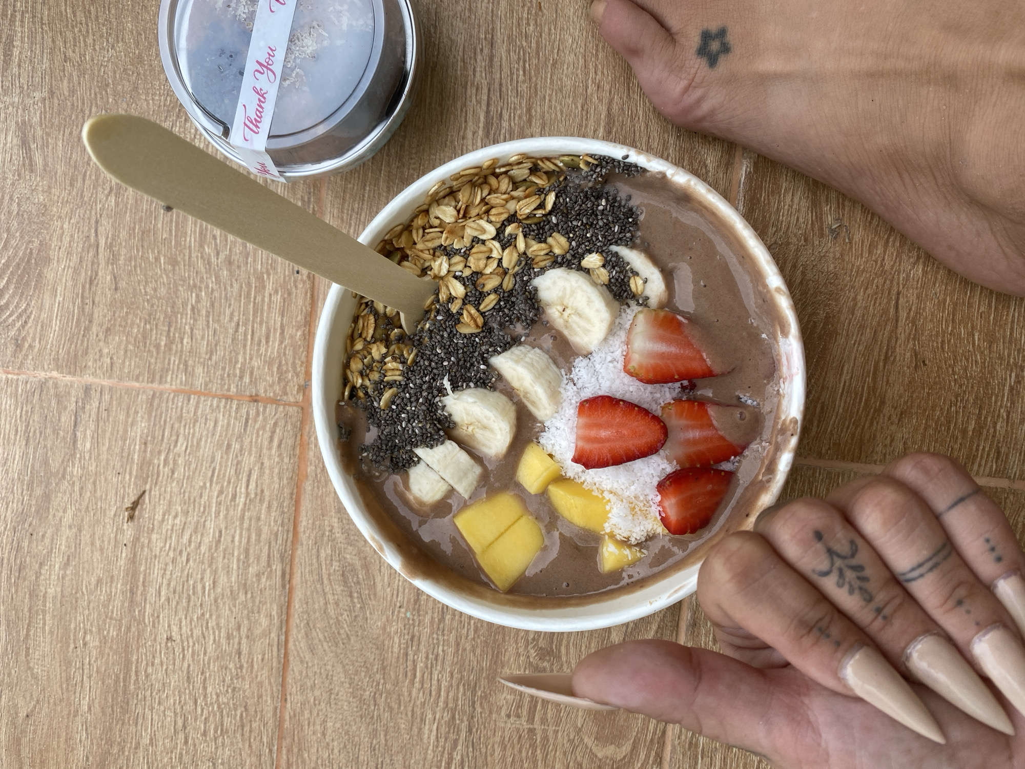 A close-up, top-down view of a chocolate smoothie bowl topped with granola, chia seeds, sliced bananas, diced mango, shredded coconut, and sliced strawberries. The bowl is white and is resting on a light brown wooden surface, likely a table or floor with wood grain. To the right of the bowl, there is a clear plastic container with a silver lid, featuring a white label with pink text that reads "Thank You". A wooden spoon is inserted into the bowl, angled upwards. In the lower left quadrant of the image, a person's left hand is visible, with long, almond-shaped beige nails and several small finger tattoos. In the lower right quadrant, a person's right foot is visible, with a small star-shaped tattoo on the top of the foot. The overall scene suggests a casual meal or snack, possibly enjoyed outdoors. The wooden surface and the warm lighting could indicate an outdoor setting in a tropical or warm climate, such as a cafe or resort in Mũi Né, Vietnam, which is known for its beaches and resorts with wooden elements.