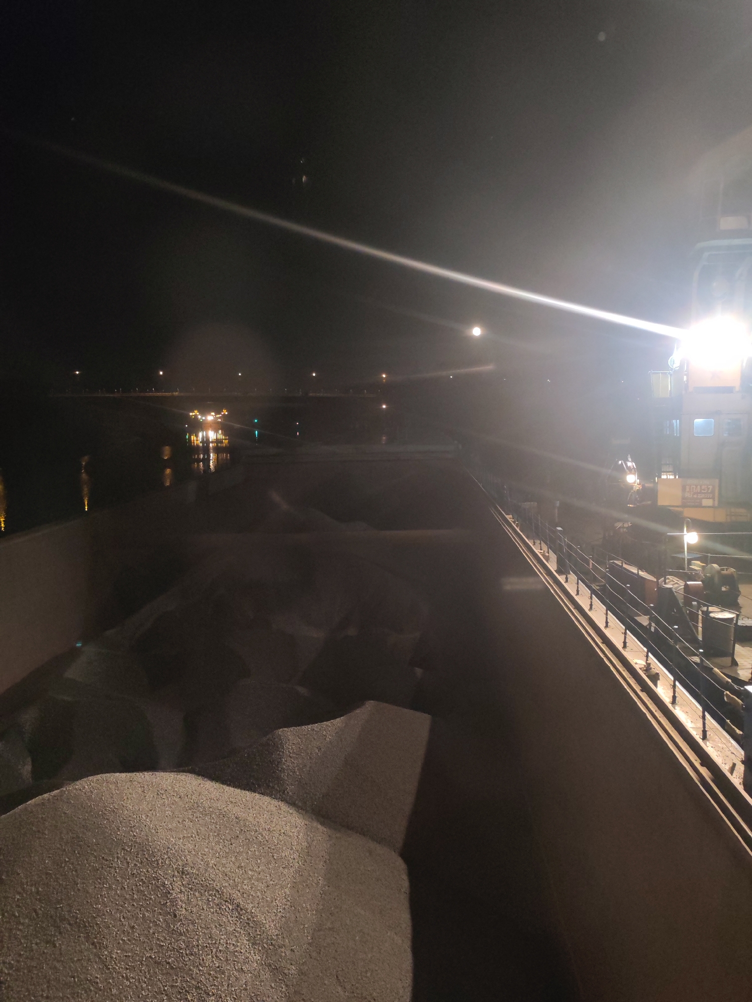 The image depicts a night scene on what appears to be a barge loaded with gravel. The scene is illuminated by bright lights emanating from the barge itself and possibly from the shore. The gravel pile occupies a large portion of the frame in the foreground. In the background, across the water, lights suggest a shoreline or a structure such as a bridge, and the reflection of these lights is visible in the water. The barge is equipped with a railing and a structure that contains what seems to be an office or control room, marked with a number, likely 10457. There are no visible people in the immediate view. It is not possible to confirm the location as Yakhroma, Russia, from the given image.