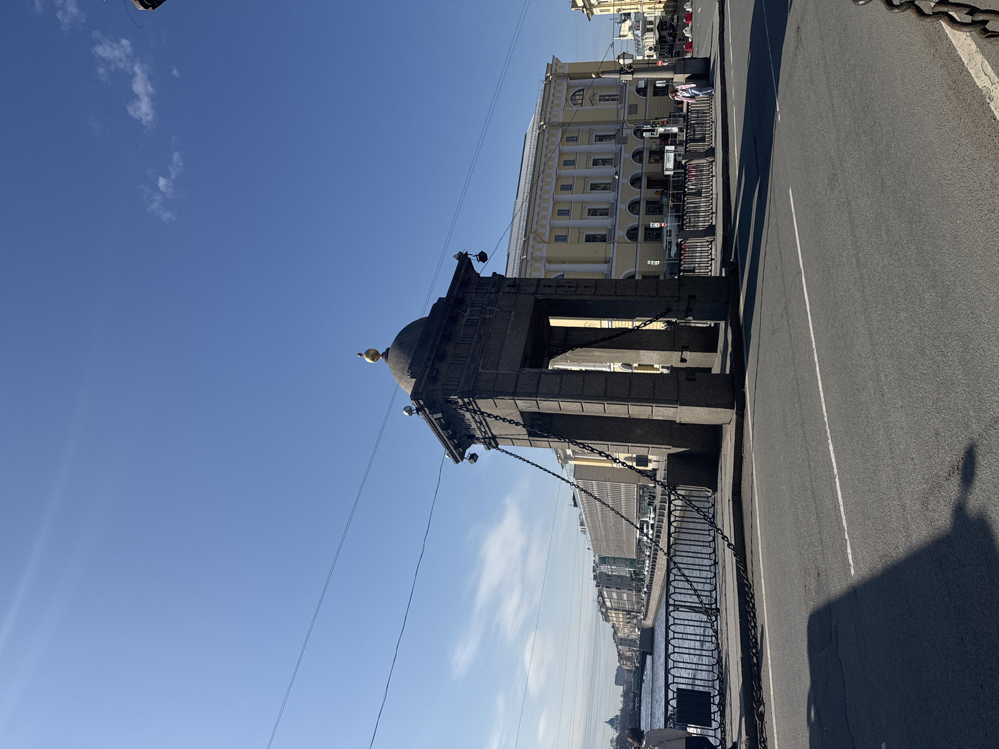 The image captures a scene in Saint Petersburg, Russia, depicting the Lomonosov Bridge and the Fontanka River. A granite pavilion atop the bridge, adorned with a dome and chains, is visible on the left side of the frame. Behind the bridge, along the riverbank, a yellow building with white columns and arched windows can be seen.
The foreground is dominated by the asphalt road of the bridge, with white lane markings. A metal fence runs along the river's edge, connected to the bridge's structure by chains.
The sky is clear and blue, with a few wisps of clouds.
The presence of the Fontanka River, the Lomonosov Bridge's distinctive architecture, and the yellow building typical of Saint Petersburg's architecture solidify the location.