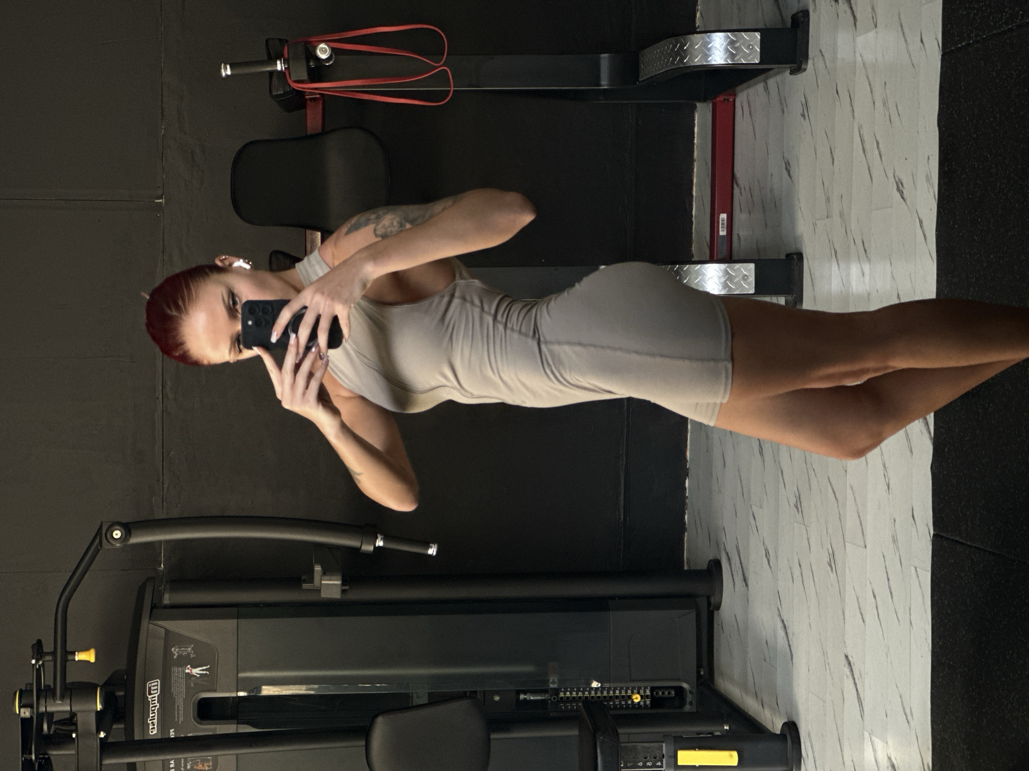 A young woman stands in a gym setting, taking a selfie with her phone. She has short red hair, fair skin, and a tattoo visible on her left arm. She is wearing a form-fitting, light-colored dress. Her face is partially obscured by her phone as she holds it up to take the picture.
The background features standard gym equipment: a black machine with yellow accents, and another piece of equipment with black and red elements, including resistance bands. The walls are a dark, possibly black, color, contrasting with the white and grey marbled flooring pattern. The scene is located in North Kuta, Indonesia, suggested by the tropical climate and gym style.