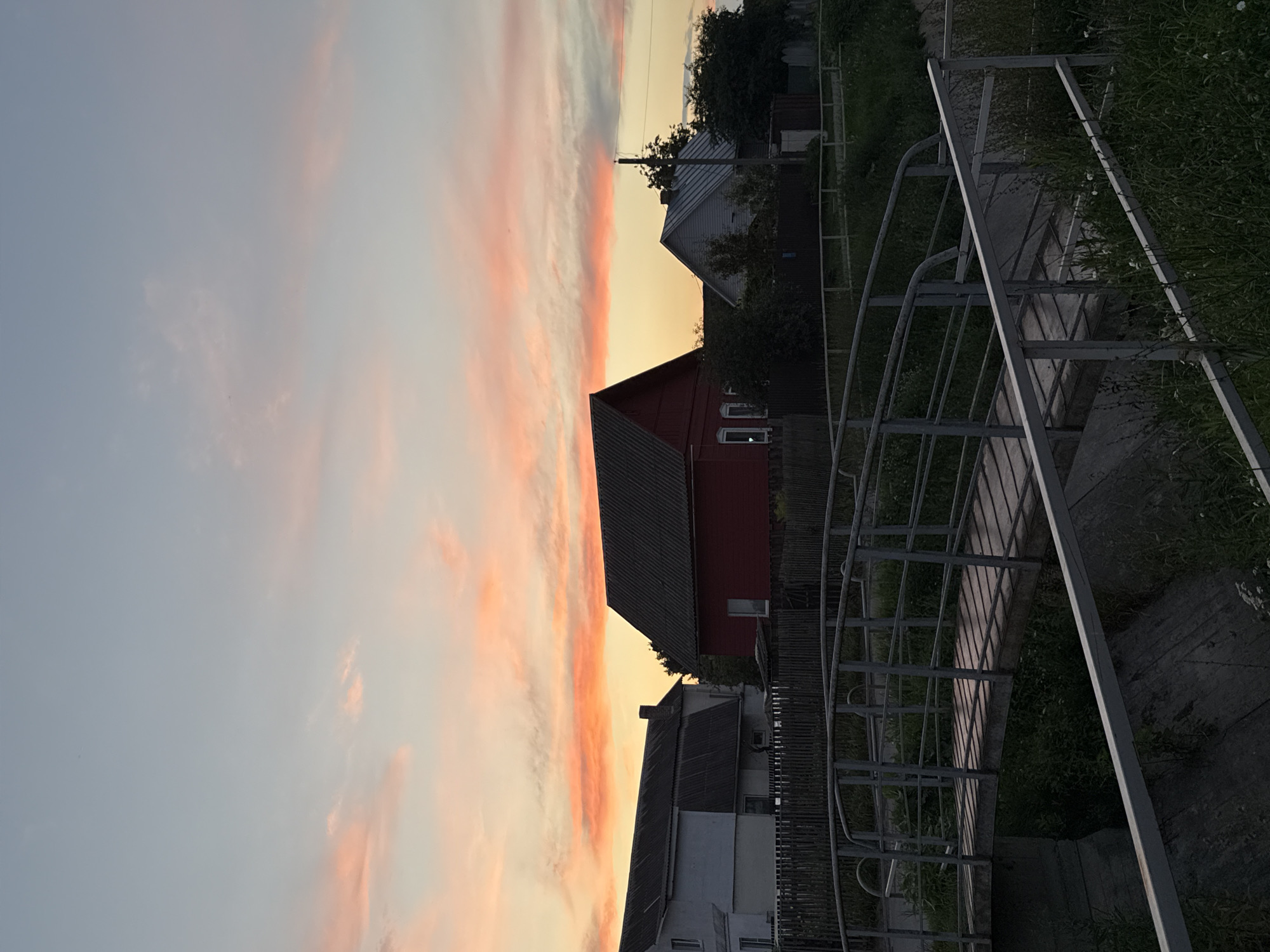 The image depicts a serene rural or suburban landscape at dusk, under a sky painted with vibrant orange and pink hues from the setting sun. In the foreground, a metal and wooden footbridge spans a small channel, with concrete structures visible below and overgrown green grass along the banks. Beyond the bridge, a row of houses with dark roofs can be seen, including a striking red house and a lighter colored building. A wooden fence separates the properties from the foreground. Trees and foliage are scattered throughout the scene, adding to the natural setting. The overall atmosphere is peaceful and quiet. The location is identified as the city of Mahilyow, Belarus.