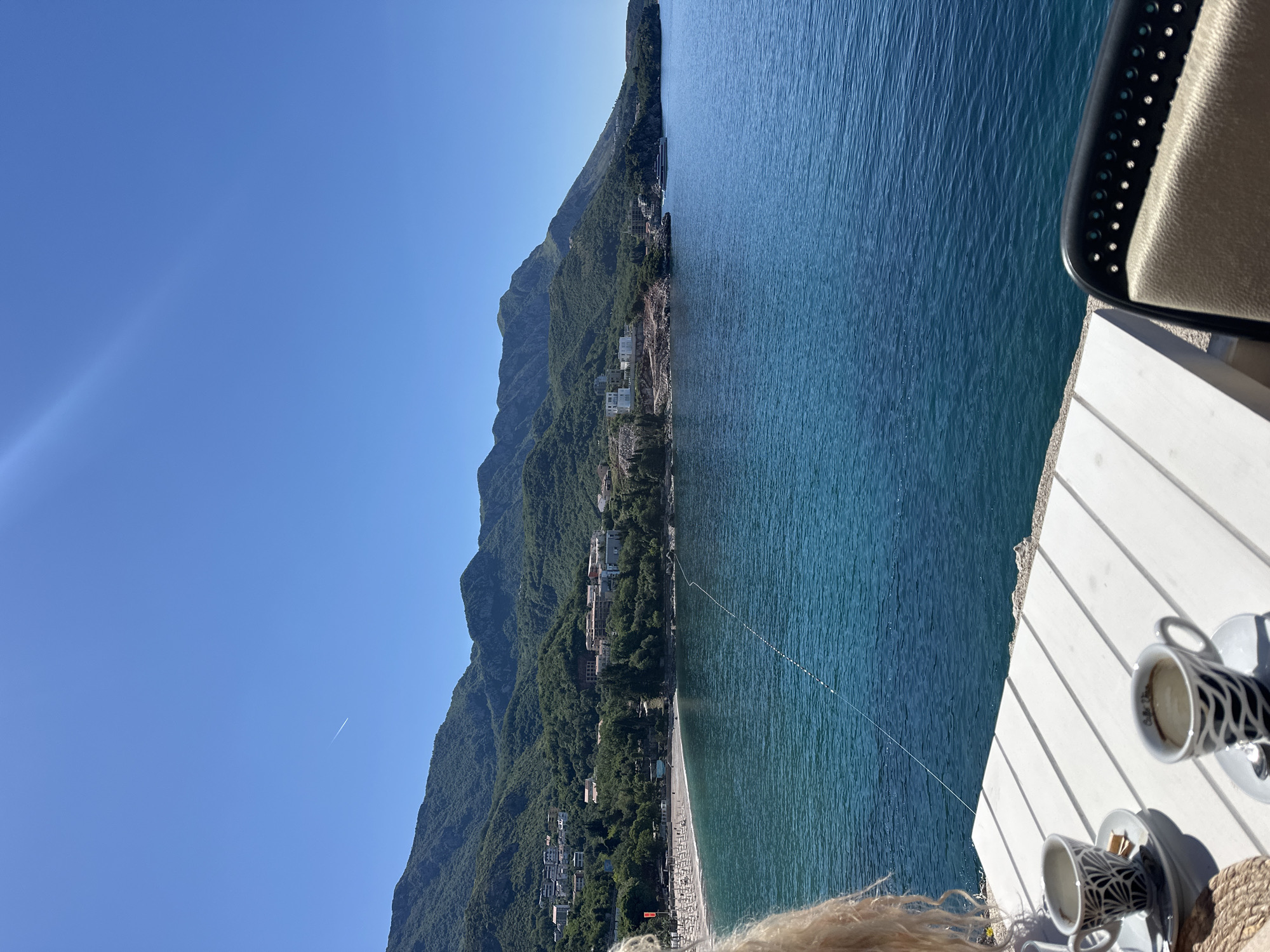 The photograph depicts a coastal scene in Boreti, Montenegro. A mountain range covered in dense greenery runs along the left side of the frame. The Adriatic Sea occupies the right side of the frame, with varying shades of blue reflecting the sunlight. A thin strip of land borders the sea, featuring structures and foliage interspersed along the coastline.
In the foreground, a white wooden table is visible, with two coffee cups and saucers placed on it. Part of a chair with a light-colored seat is seen at the top right corner. At the bottom, blonde hair is visible. The sky above is clear blue, with a contrail from an airplane.
