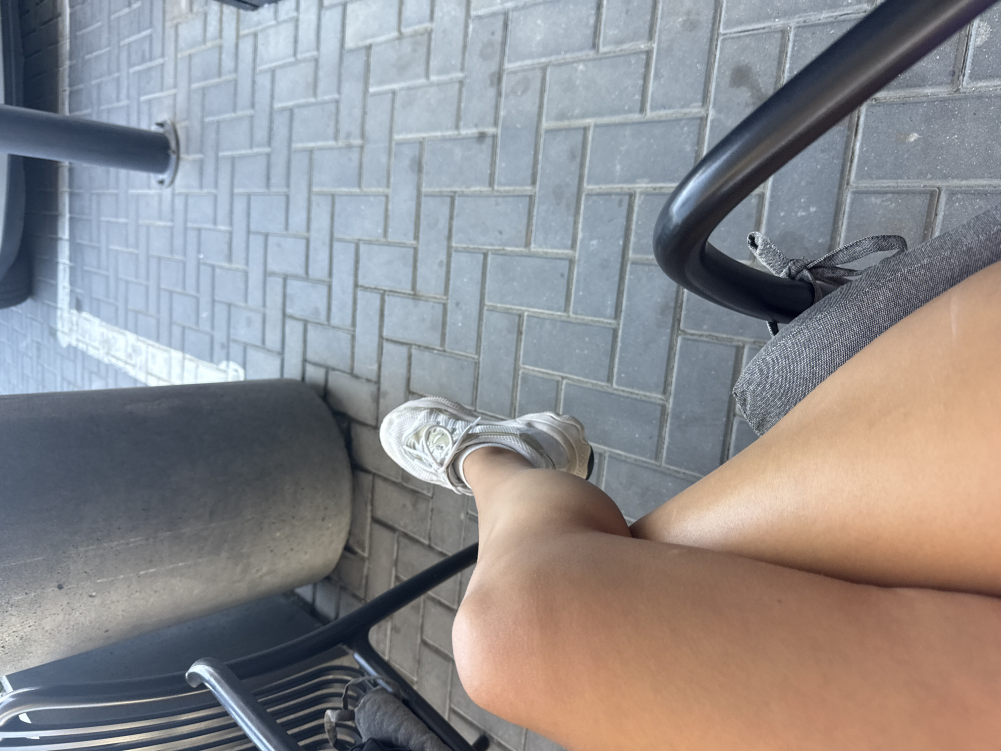 A person's legs, wearing white sneakers, are crossed while sitting on a black metal chair with a gray fabric seat. The person is outdoors, with a grey, textured concrete planter and a grey brick pavement visible in the background. The scene is set in Comrat, Moldova.