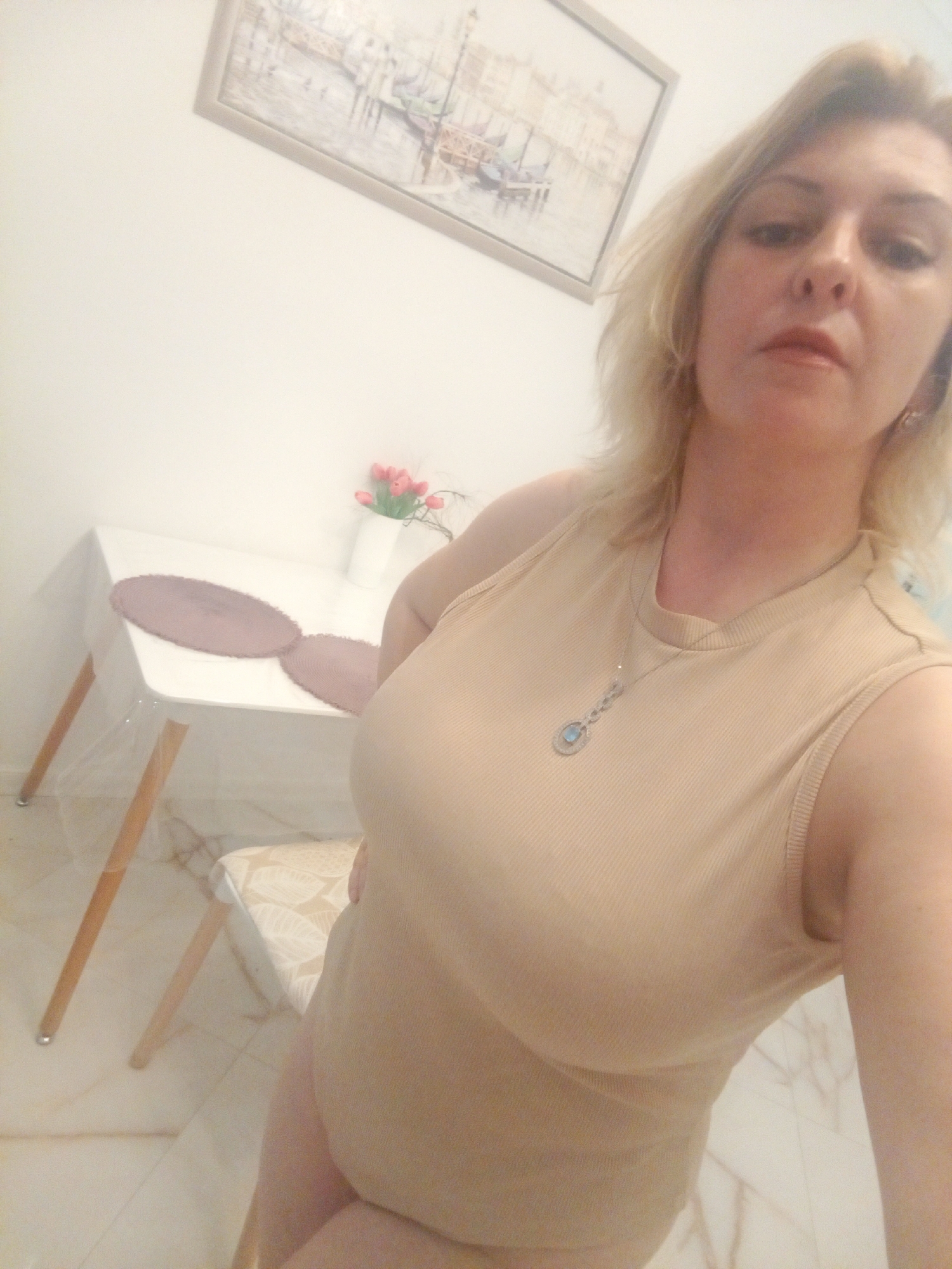 The media file shows a woman with blonde hair and fair skin, wearing a light beige tank top and a necklace. She is taking a selfie in what appears to be an indoor setting, possibly a dining area or living space. Behind her, a small white table is set with two oval placemats and a white vase containing pink tulips. A framed picture hangs on the wall above the table, depicting a scene with buildings and boats. The flooring appears to be tiled with a marble-like pattern. The lighting suggests it could be daytime, as there is no indication of artificial lighting. The woman's expression is neutral. There is no visible text in the image. The context provided states the location is Sochi, Russia.