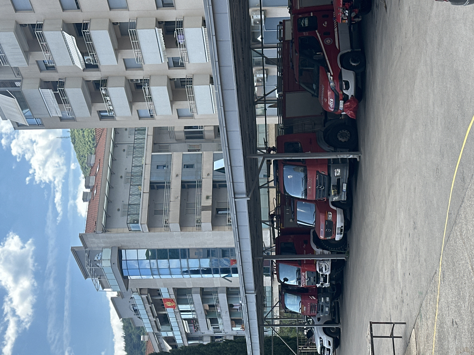 The image depicts a fire station in Budva, Montenegro. The station houses a row of fire trucks under a covered parking structure. Adjacent to the fire station is a modern, multi-story building with glass balconies. The sky above shows scattered clouds.