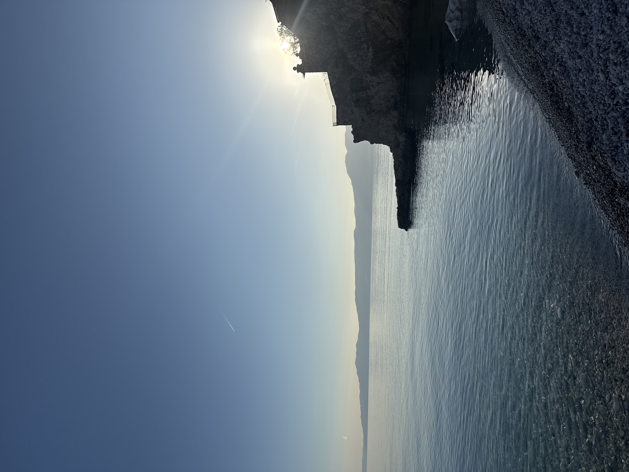 The image shows a coastline in Grad Rijeka, Croatia. The scene includes a rocky cliff face on the right side, a narrow beach with pebbles or small rocks, and calm blue water extending towards the horizon. In the background, there are other landmasses or islands visible. A building or structure is visible on top of the cliff. The sun is shining brightly behind the cliff, creating a lens flare effect. A contrail from a jet is visible across the sky.