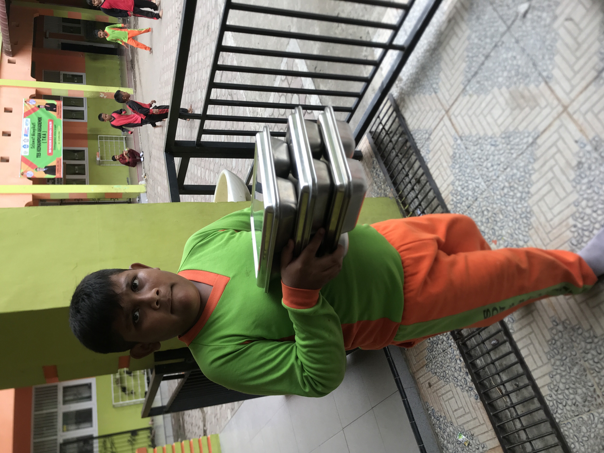 A young boy in a green and orange uniform stands outside, holding a stack of silver metal trays. He is positioned next to a black metal railing and patterned gray tiles. In the background, other children, also in similar uniforms, are visible near a building with a banner in Indonesian. The location is in Lhokseumawe, Indonesia. The building is painted in shades of orange, green, and yellow.