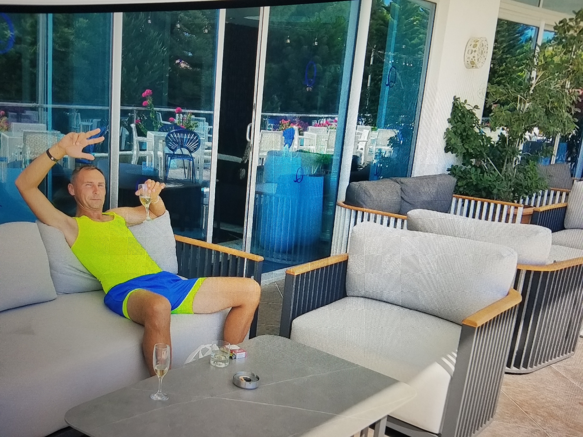 A man with short, light-colored hair and a light complexion is sitting on a light-gray sofa. He is wearing a neon yellow tank top and blue shorts with a yellow trim. He is holding a glass of champagne in his right hand and has his left arm raised, with his hand open, fingers spread. He is looking towards the camera with a slight smile. In front of him, on a light-gray coffee table, are another glass of champagne, a small packet of cigarettes, and an ashtray. The sofa and table are on an outdoor patio area with light-gray cushioned chairs and wooden railings visible in the background. Behind the patio area, there are large glass doors that appear to look into a restaurant or bar area with tables and chairs. The environment suggests a relaxed, possibly vacation-like setting. The image does not provide any specific visual cues that would place the location in Tiraspol, Moldova.