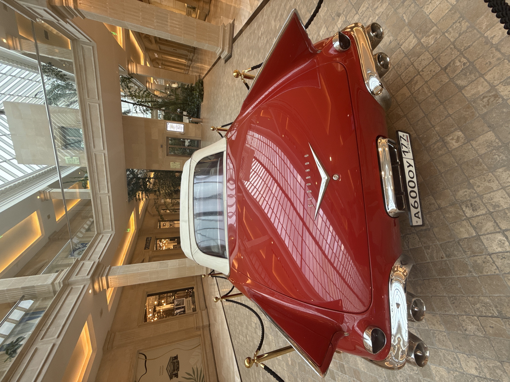 An overhead shot displays a classic red convertible car with a white top, parked indoors. The car has a prominent chrome grille and distinctive tail fins. Its license plate reads "A600OY 177 RUS". The car is cordoned off with black ropes and gold stanchions, suggesting it's an exhibit or display.
The setting appears to be a spacious, modern atrium with a high, glass-paneled ceiling that reflects the car's polished surface. Tall, light-colored columns and arched doorways line the perimeter, hinting at a shopping mall, hotel lobby, or a similar public indoor space. The floor is paved with textured stone tiles. Artificial lighting illuminates the area, with warm light coming from recessed ceiling fixtures and shopfronts visible in the background. Some greenery, possibly potted plants, can be seen in the background.
The image is well-lit, and there are no people visible in the frame. The overall impression is one of elegance and display. The time of day and weather are not discernible, but the indoor setting implies controlled conditions. The visible text includes the license plate and potentially some signage in the background, but it is too small and blurry to be read.