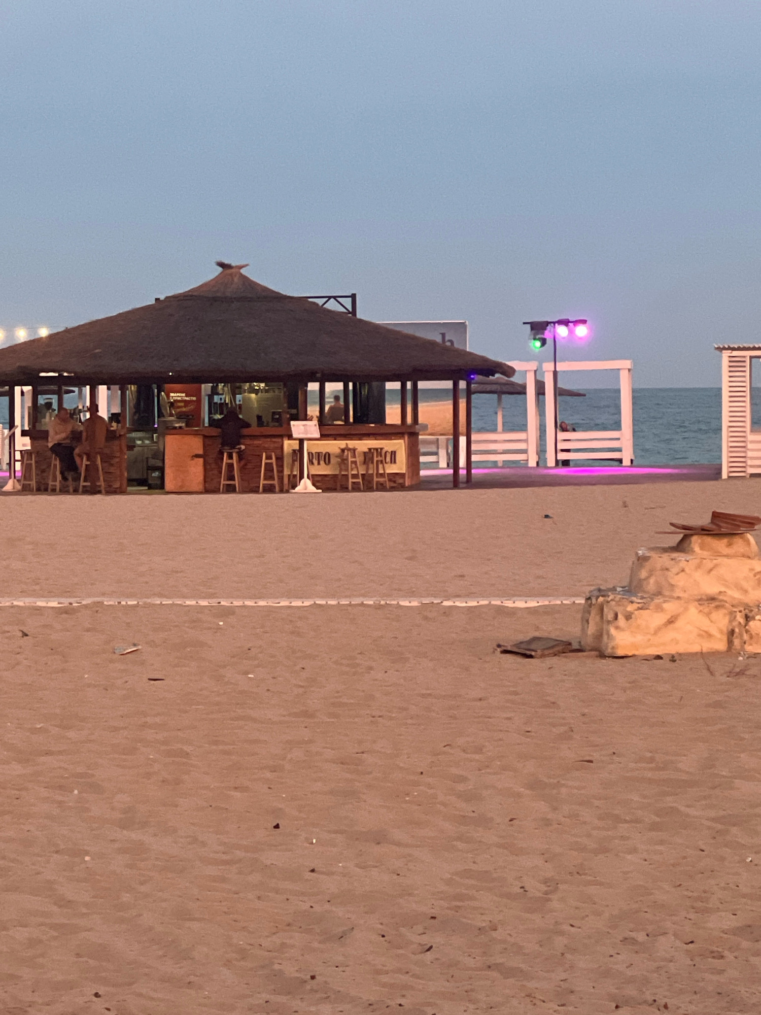 The image captures a beachside scene at dusk in Zatoka, Ukraine, likely near the Black Sea. In the foreground, a sandy beach stretches towards a thatched-roof bar with several patrons seated on stools. The bar appears to be made of wood and stone. In the background, illuminated by colored stage lights, are white structures and the sea. The sky is a soft, dusky blue. The overall impression is one of a relaxed evening atmosphere at a coastal establishment.
