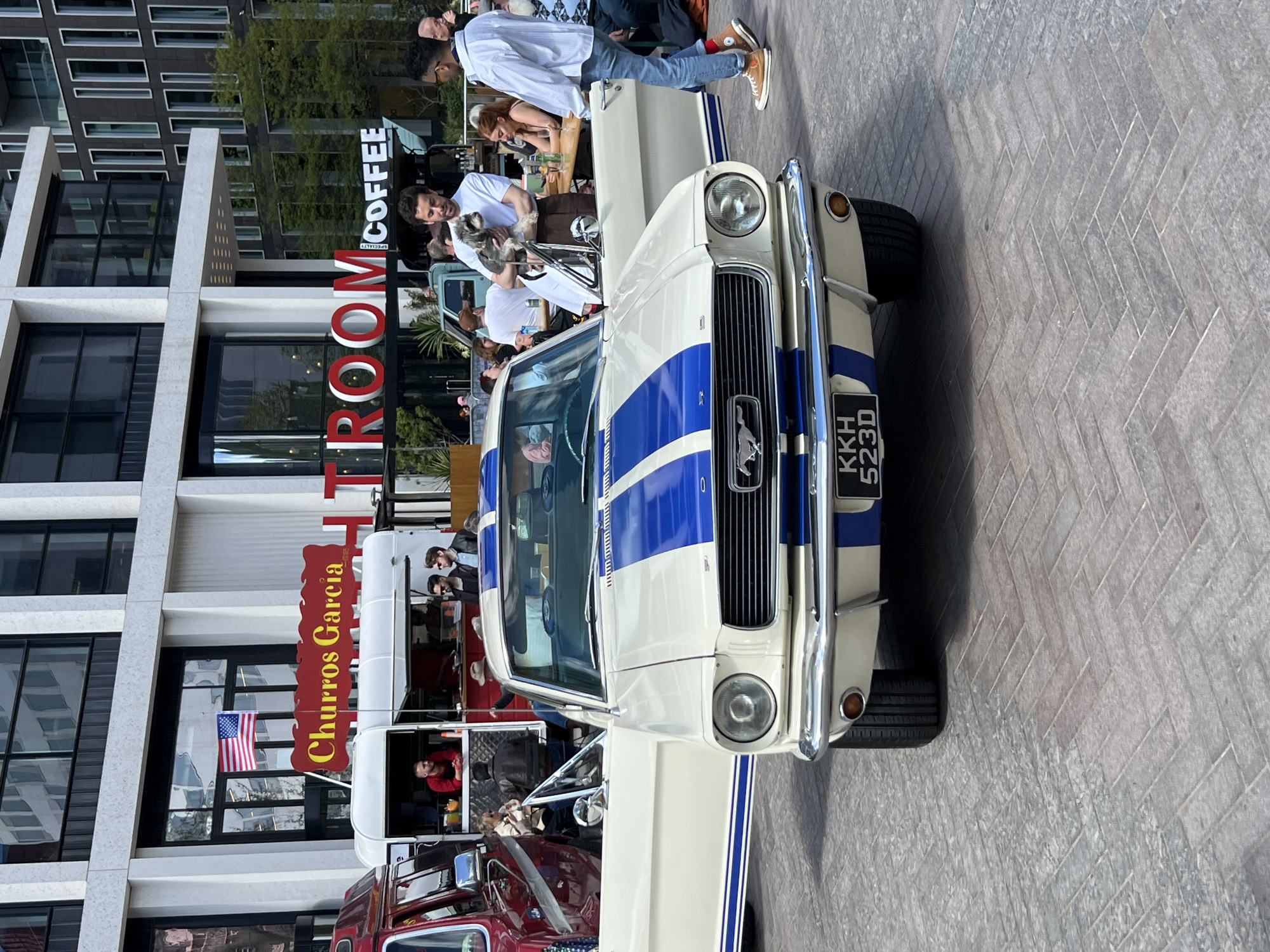 The image shows a white and blue vintage Ford Mustang parked on a paved area in what appears to be a city setting. The car has a British license plate "KKH 523D". In the background, there is a modern building with large windows and a sign that reads "ROOM Specialty COFFEE". A "Churros Garcia" food truck is also visible behind the Mustang, with people standing and sitting near the truck, possibly customers. Several people are standing around the Mustang as well. An American flag is hanging in the window behind the truck. The location appears to be a commercial area with shops and dining establishments. Based on the license plate of the Ford Mustang and the buildings visible, the location is most likely in London, United Kingdom.