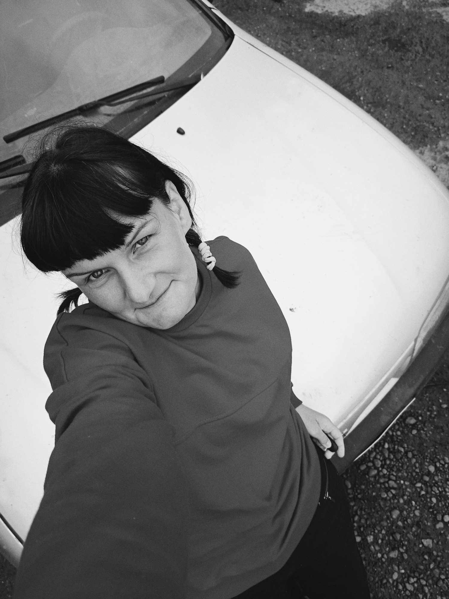 This black and white, high-angle image captures an adult female situated in front of a light-colored vehicle, likely a car, in what appears to be an unpaved outdoor setting in Большая Уса, Russia.
The subject is a female with dark hair styled with bangs across her forehead and two small pigtails secured with light-toned hair ties, one visible on the right side of her head. She faces the camera directly, her head tilted slightly, with a subtle smirk or gentle smile. Her eyes are open, and her gaze is directed towards the lens. She is wearing a dark-toned, long-sleeved garment, possibly a sweatshirt, which covers her torso and arms. Her left hand is partially visible near her waist, resting on an object that appears to be the front edge of the car's hood or bumper, with a zipper pull visible on her lower garment.
The vehicle occupies a significant portion of the frame, predominantly the upper right and lower sections. It is a light-colored car, appearing white or a pale tone in the grayscale rendition. The front hood, windshield, and windshield wipers are clearly visible in the upper half of the image. The wipers are positioned horizontally across the base of the windshield. The very front bumper or lower grille area is visible along the bottom edge of the frame. The car's surface shows some minor smudges or dust, suggesting moderate use.
To the right and bottom right of the vehicle, the ground is visible. It is an unpaved surface composed of varying sizes of dark-toned gravel and dirt, indicating an outdoor, possibly rural or semi-urban environment. The lighting appears to be natural daylight, casting no harsh shadows, suggesting an overcast day or diffused light. The overall tone of the image is monochromatic, with a moderate range of grays.