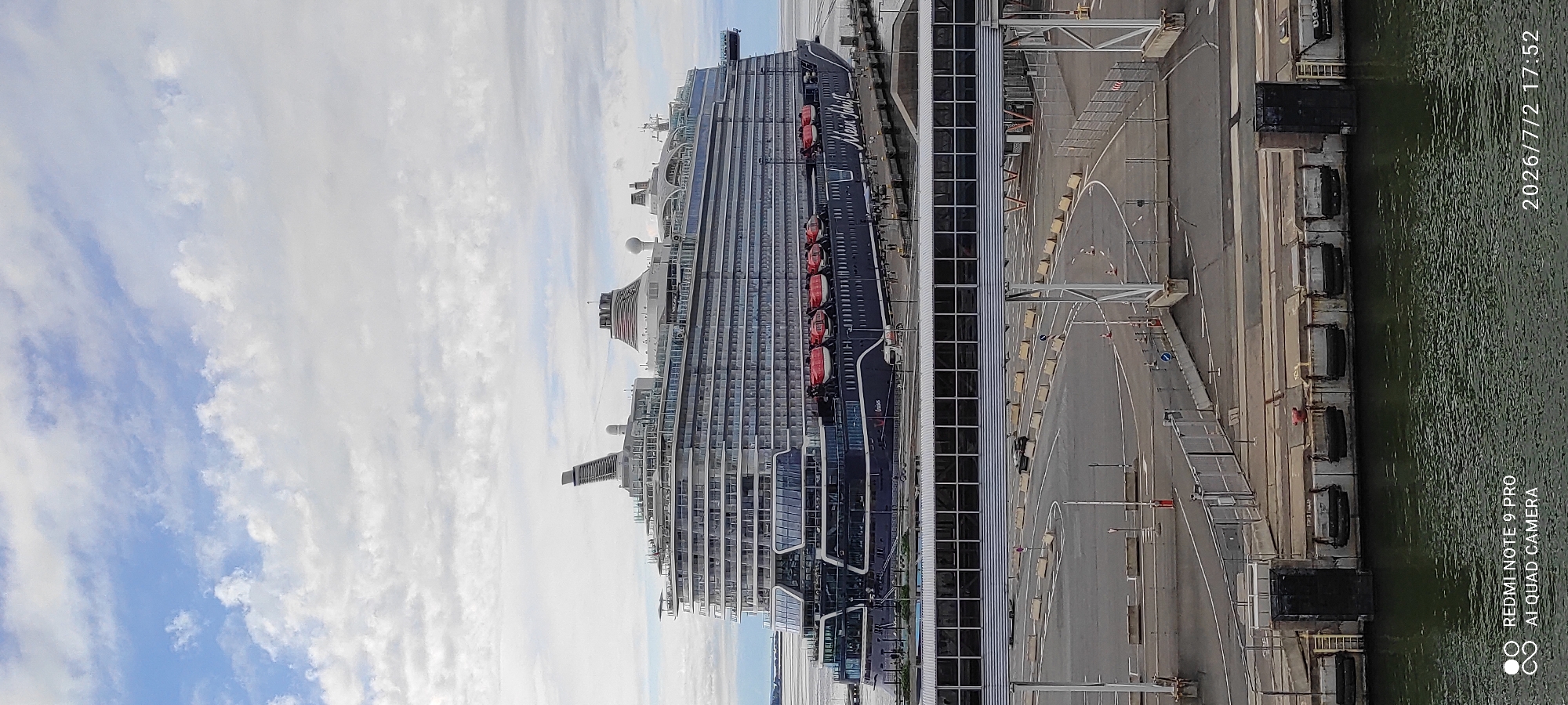 The image depicts a large cruise ship docked at a port, likely in Tallinn, Estonia, given the location context. The ship, identified by the text "Mein Schiff" on its hull, is dark blue with several decks featuring numerous windows. Orange lifeboats are visible along the side of the ship. Adjacent to the ship is a modern terminal building with large glass windows, and beyond that, a paved port area with walkways, barriers, and some parked vehicles. The water in the foreground is calm and appears greenish. The sky is partly cloudy with a mix of white and blue. The timestamp "2026/7/2 17:52" is visible in the upper right corner of the image, suggesting the time of day is late afternoon. The overall scene is one of a busy port with a massive vessel ready for or having just completed a journey. The image also contains a watermark at the bottom right corner that reads "REDMI NOTE 9 PRO AI QUAD CAMERA". No people are clearly visible in the image, contributing to a sense of calm and order at the port.